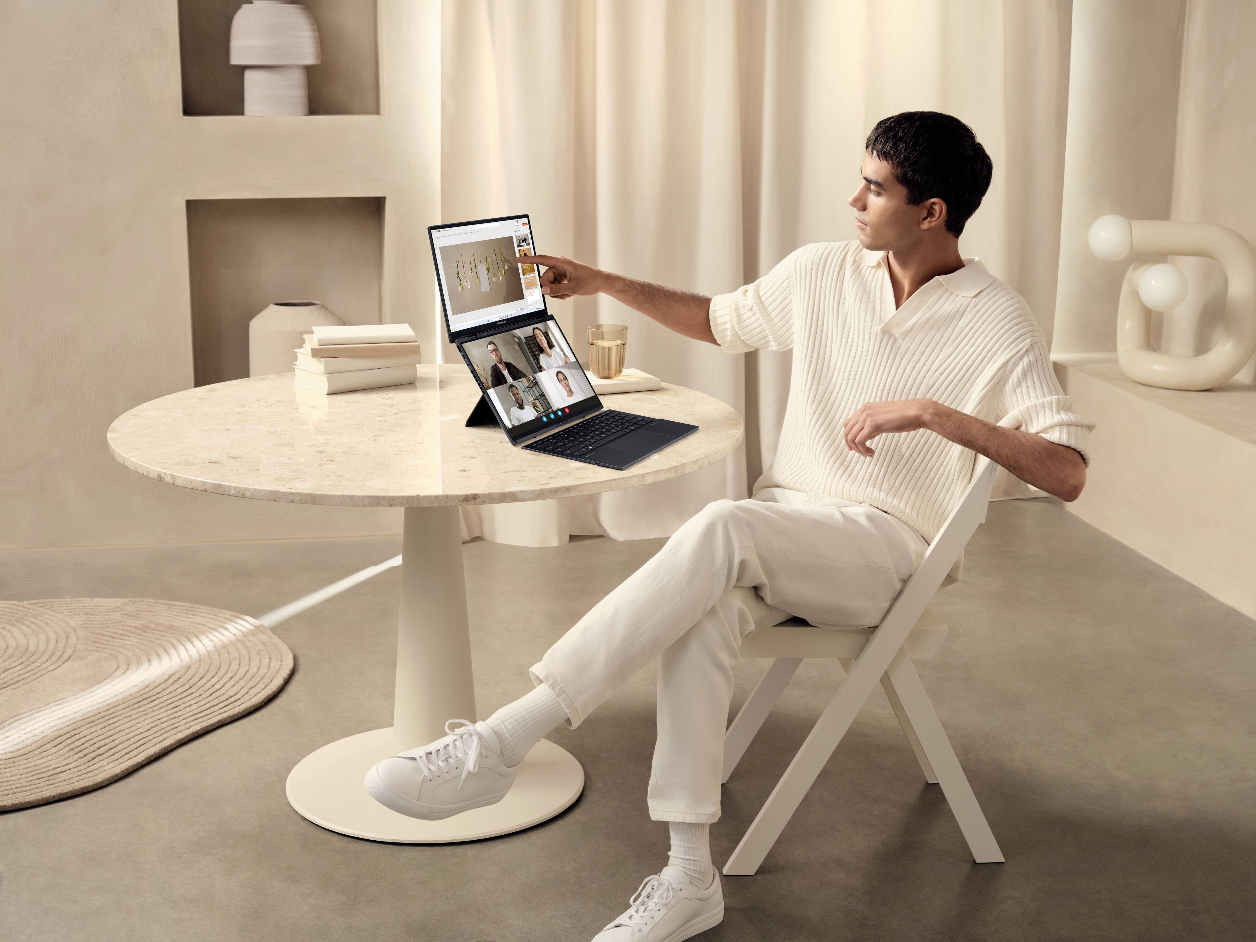 Ein Mann sitzt an einem Tisch und benutzt einen Laptop mit einem zweiten Bildschirm. Auf dem Bildschirm ist eine Videokonferenz zu sehen. Der Raum hat eine minimalistische, helle Einrichtung.