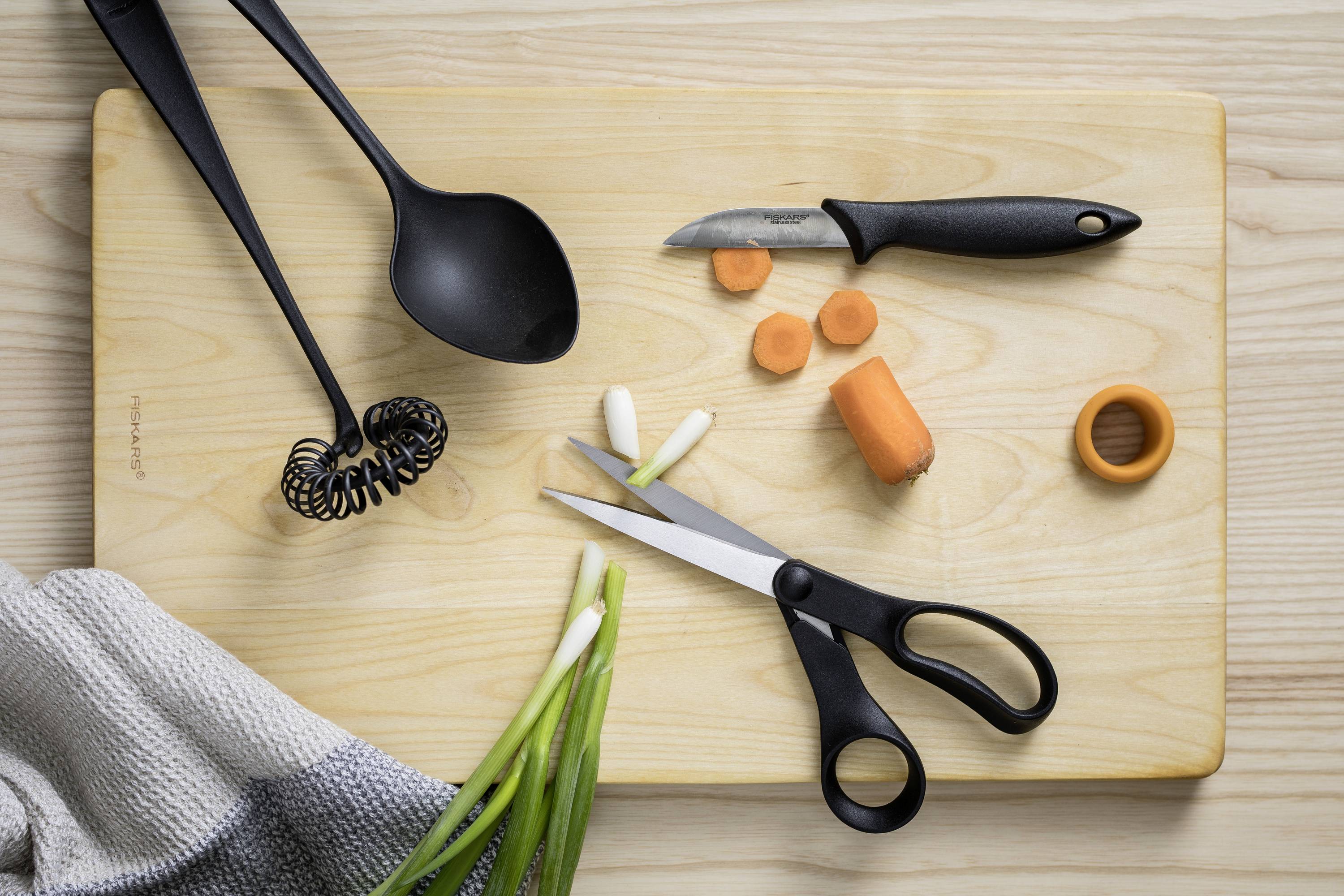Ein Schneidebrett mit Küchenutensilien: Schöpflöffel, Schäler, Schere, geschnittene Karotte und Frühlingszwiebel auf Holztisch.