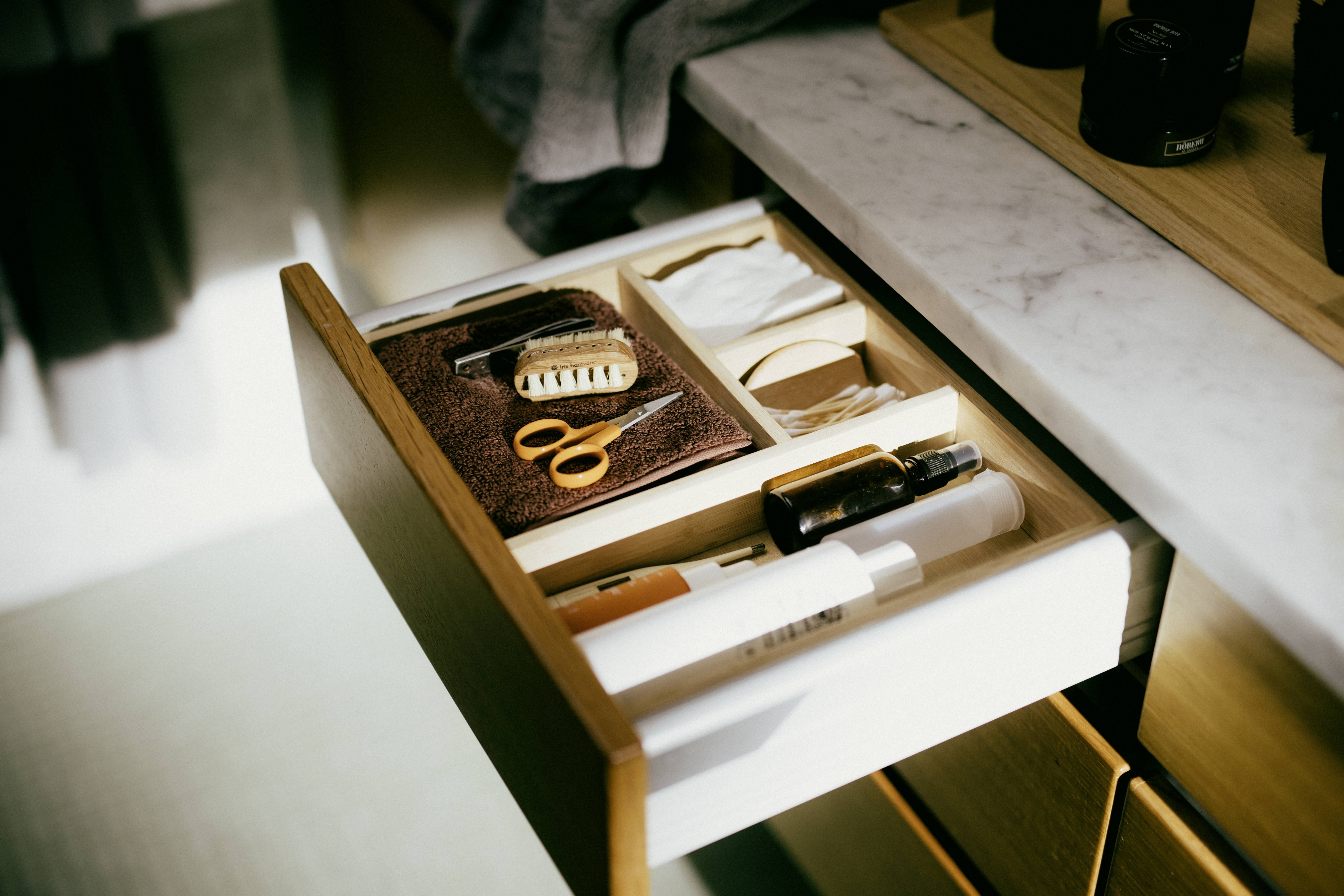 Offene Schublade mit Handtuch, Schere, Bürste und Kosmetikartikeln. An einem Waschbecken mit Marmorplatte in einem Badezimmer.