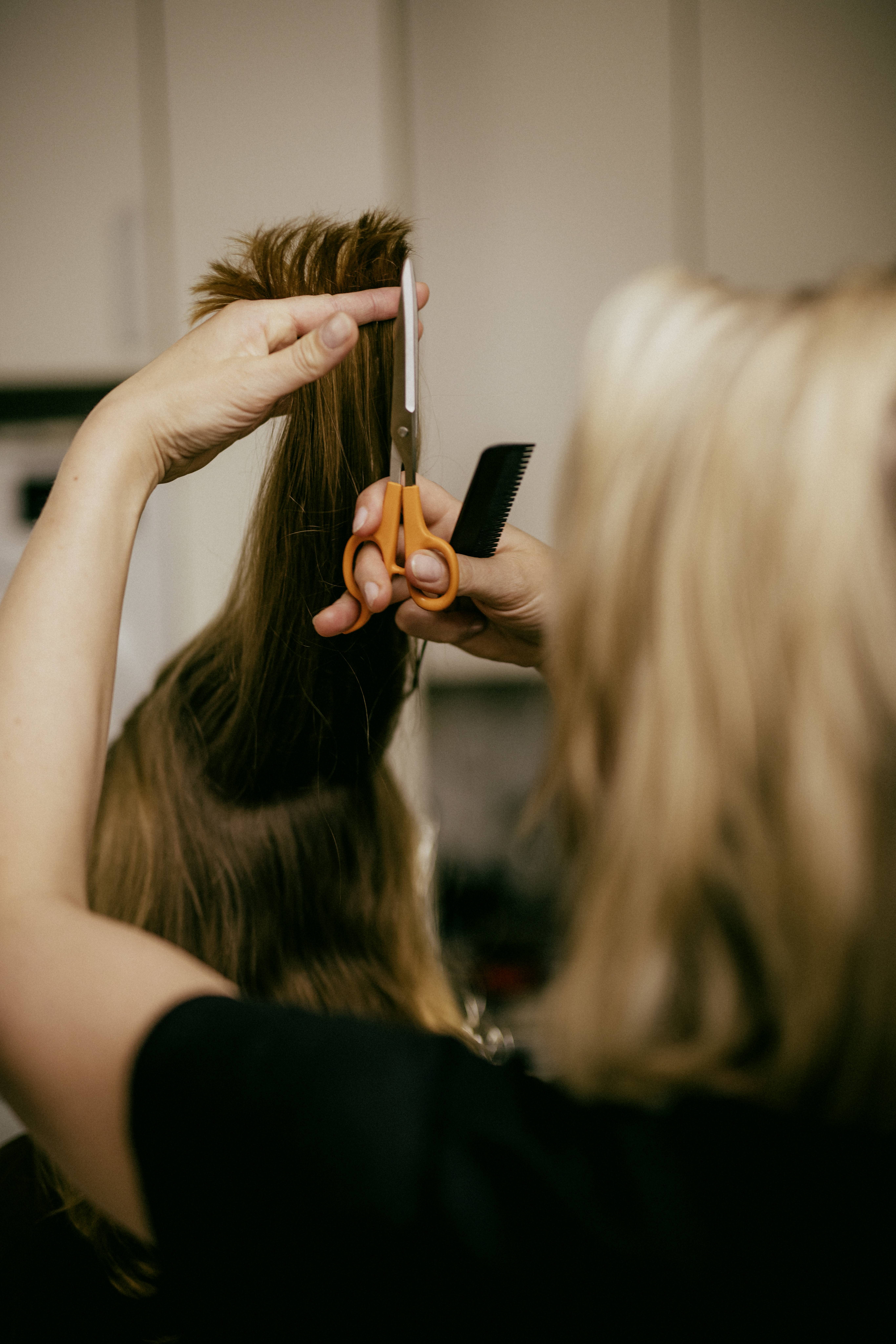Eine Person schneidet mit einer Schere das Haar einer anderen Person. Im Hintergrund sind Küchenschränke zu sehen.