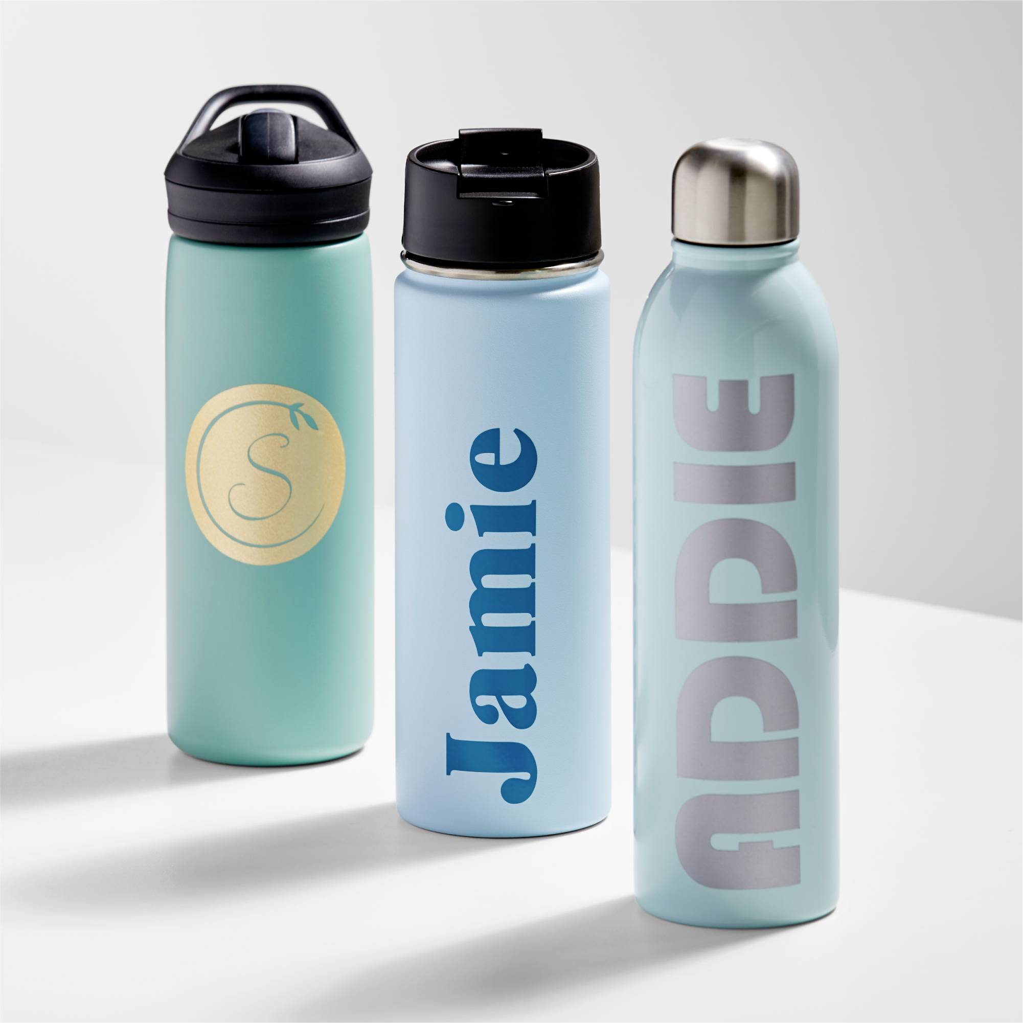 Drei Wasserflaschen nebeneinander auf einem Tisch. Eine hat ein 'S' auf einem goldenen Kreis, die anderen tragen die Namen 'Jamie' und 'ADDIE'.