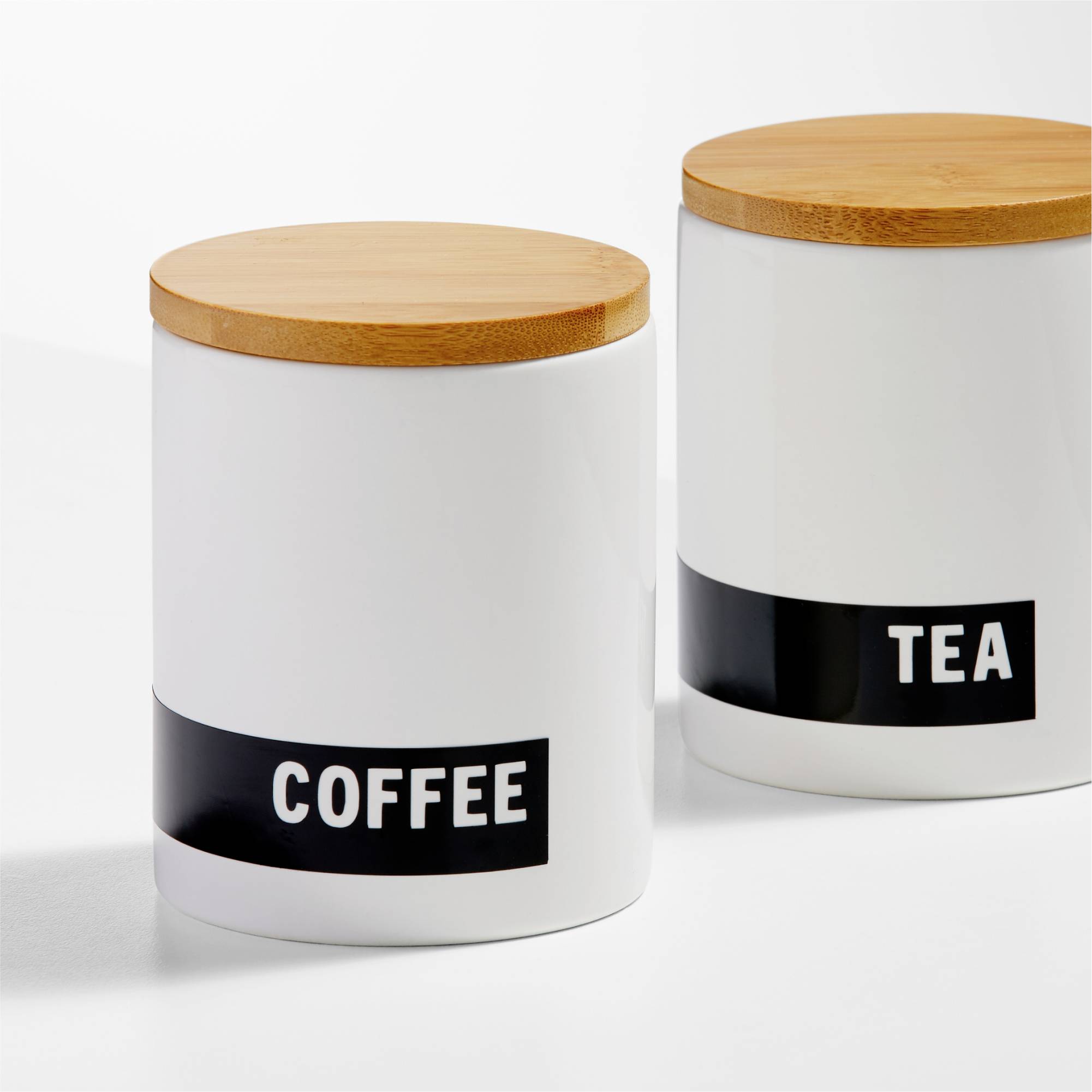 Zwei weiße Vorratsbehälter mit Holzdeckeln, mit Aufschriften 'COFFEE' und 'TEA'. Sie stehen nebeneinander auf einer weißen Oberfläche.