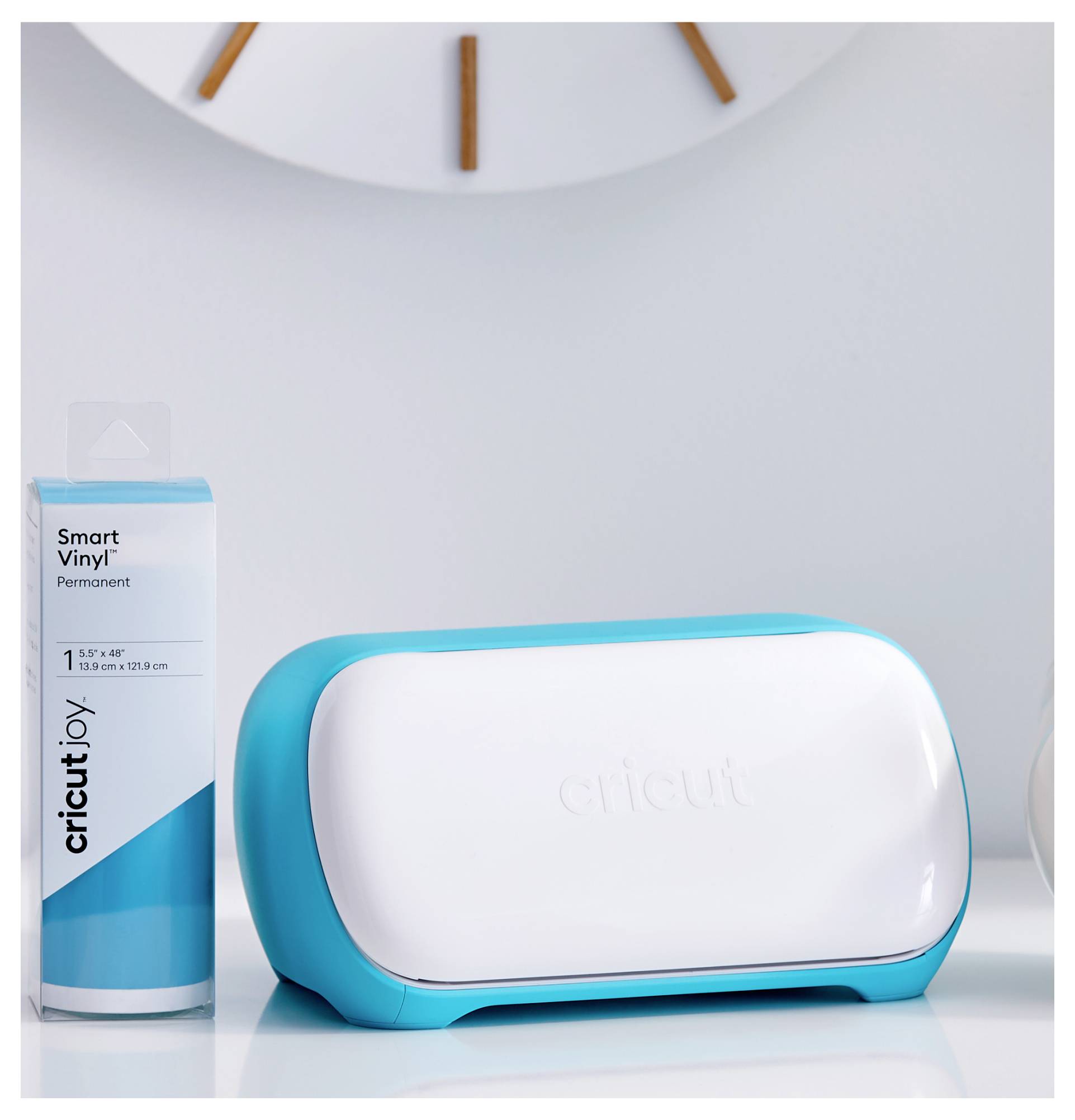Eine 'Cricut Joy'-Schneidemaschine in Blau und Weiß steht auf einem Tisch neben einer Packung Smart Vinyl, im Hintergrund eine Uhr.