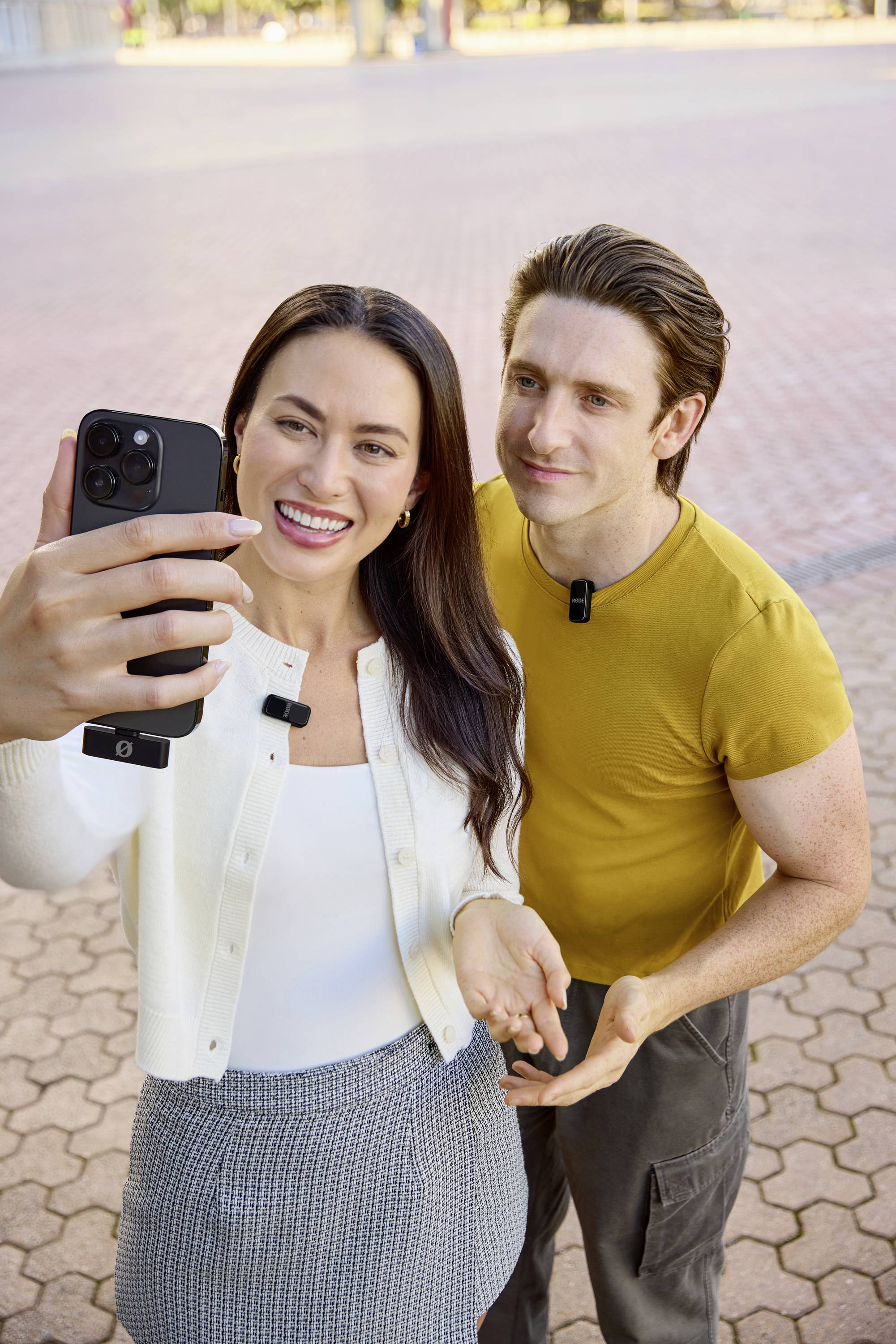 Eine Frau und ein Mann stehen draußen auf einem gepflasterten Platz, schauen fröhlich in ein Smartphone und machen ein Selfie. Beide tragen Mikrofone um den Hals.
