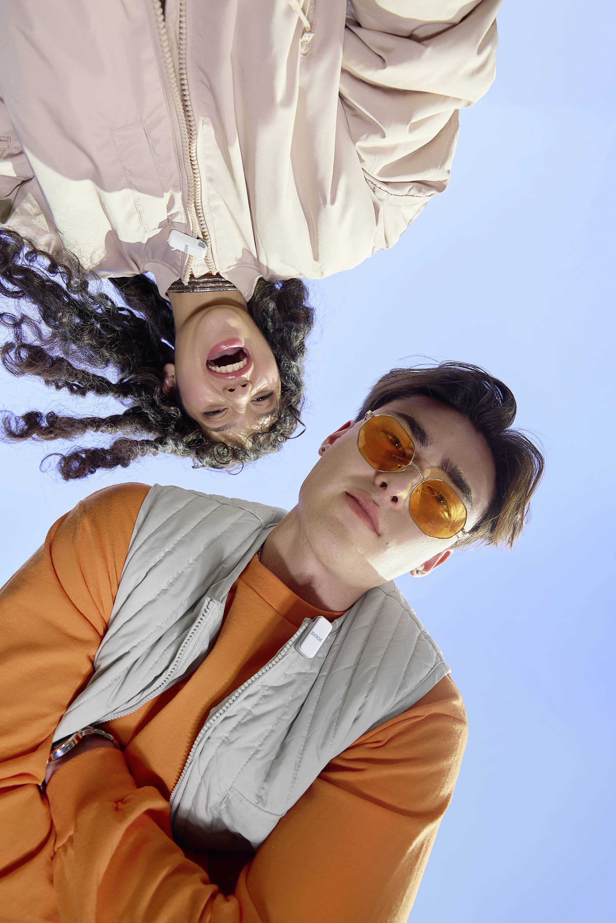 Ein Mann mit orangefarbener Sonnenbrille und eine lachende Frau mit lockigem Haar blicken in die Kamera unter freiem Himmel.