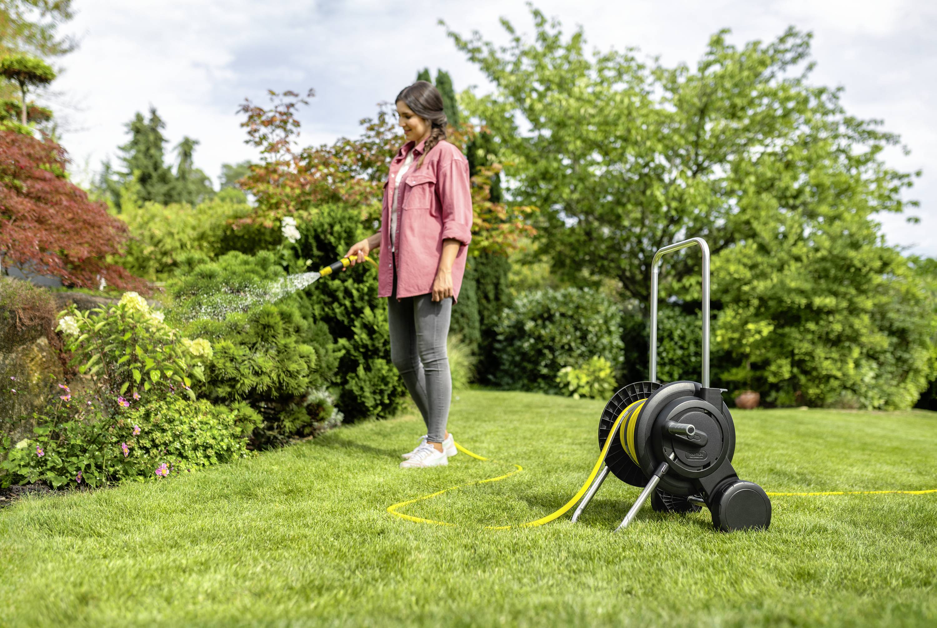 Eine Frau bewässert mit einem Gartenschlauch Blumen in einem grünen Garten. Im Hintergrund sind Büsche und Bäume zu sehen.