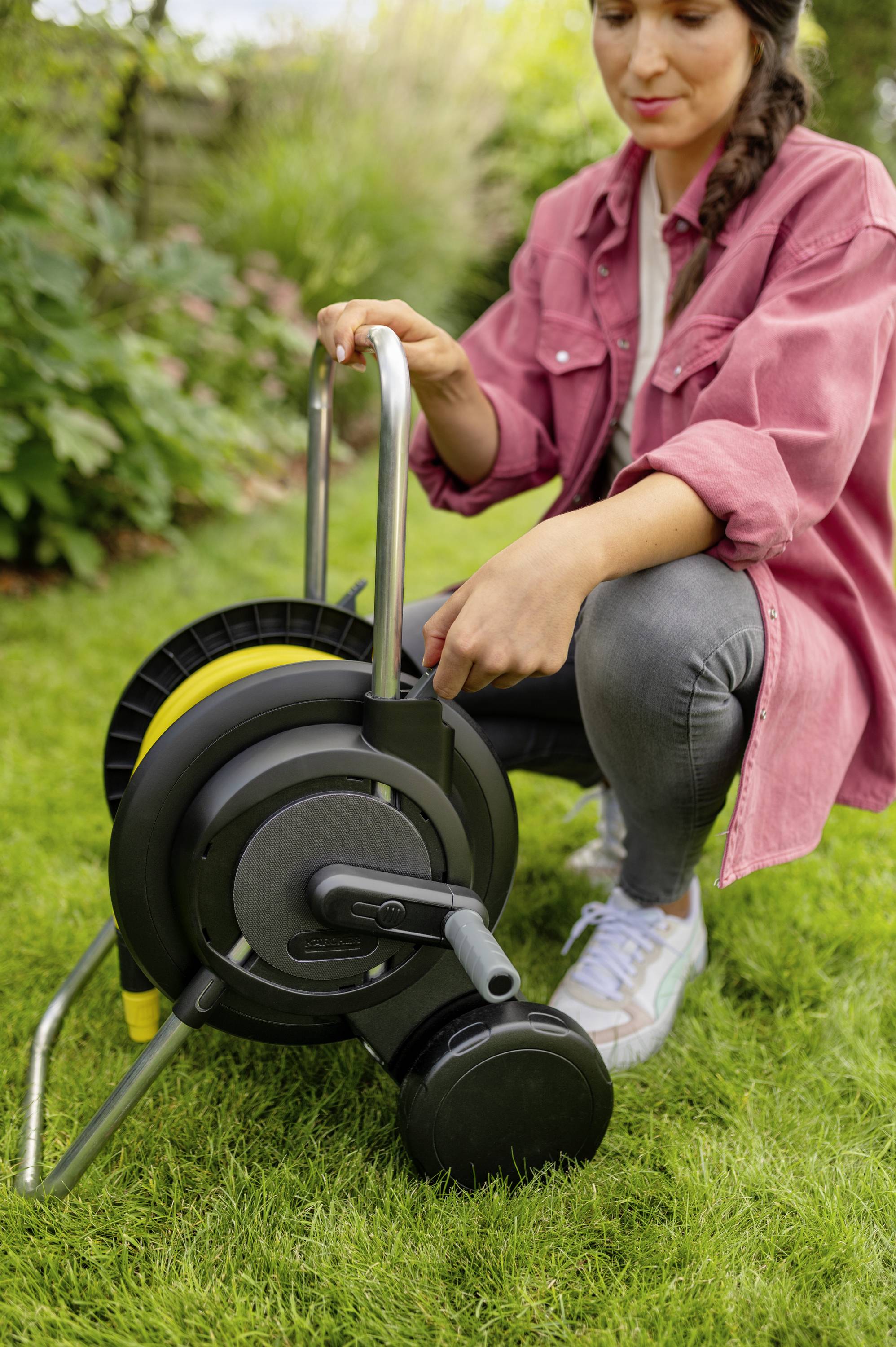 Eine Person kniet auf dem Rasen und benutzt einen Gartenschlauchwagen. Im Hintergrund sind Pflanzen zu sehen.