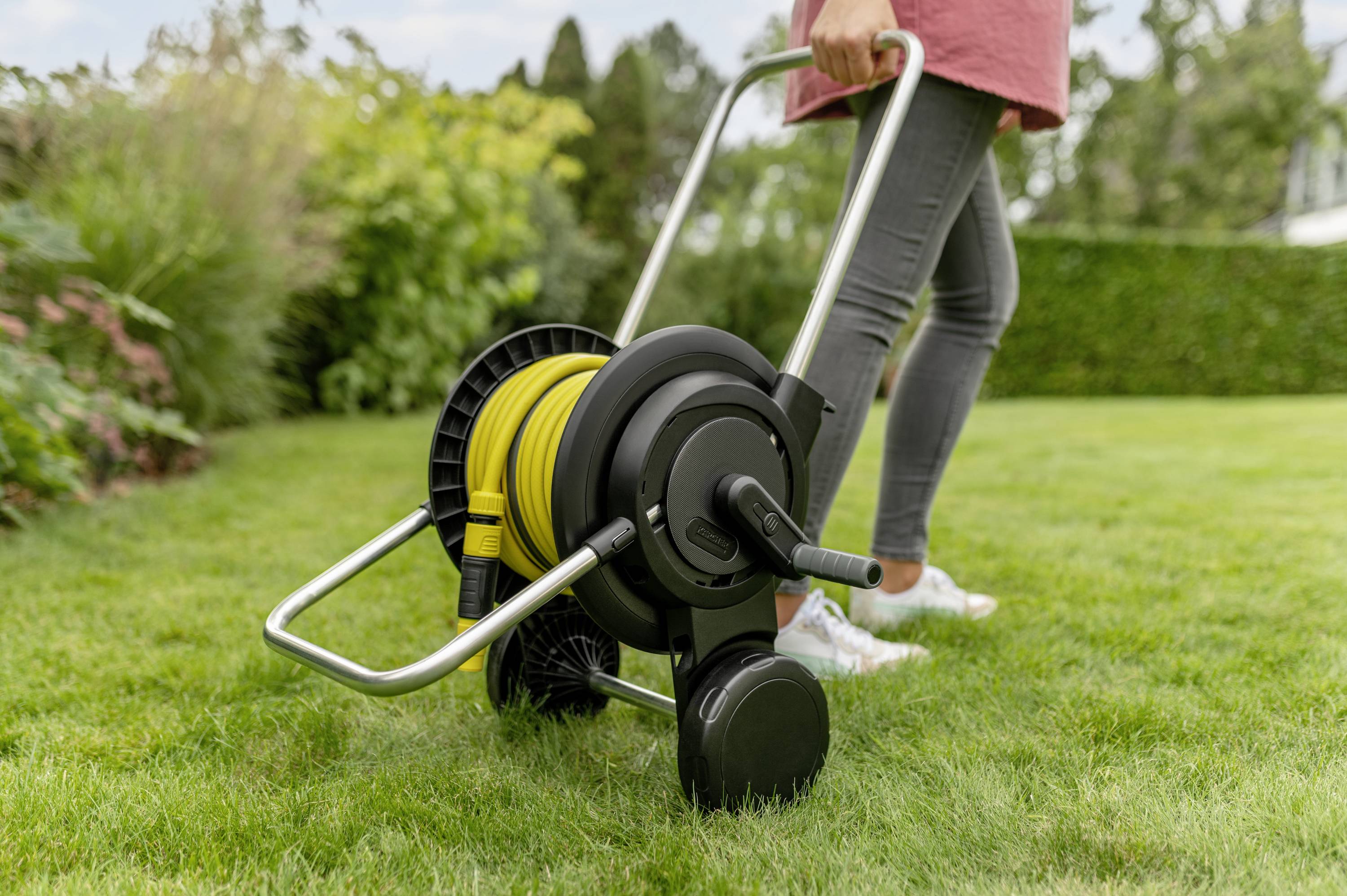 Eine Person zieht einen mobilen Gartenschlauchwagen über den Rasen. Der Schlauch ist gelb und der Wagen hat einen Metallrahmen.