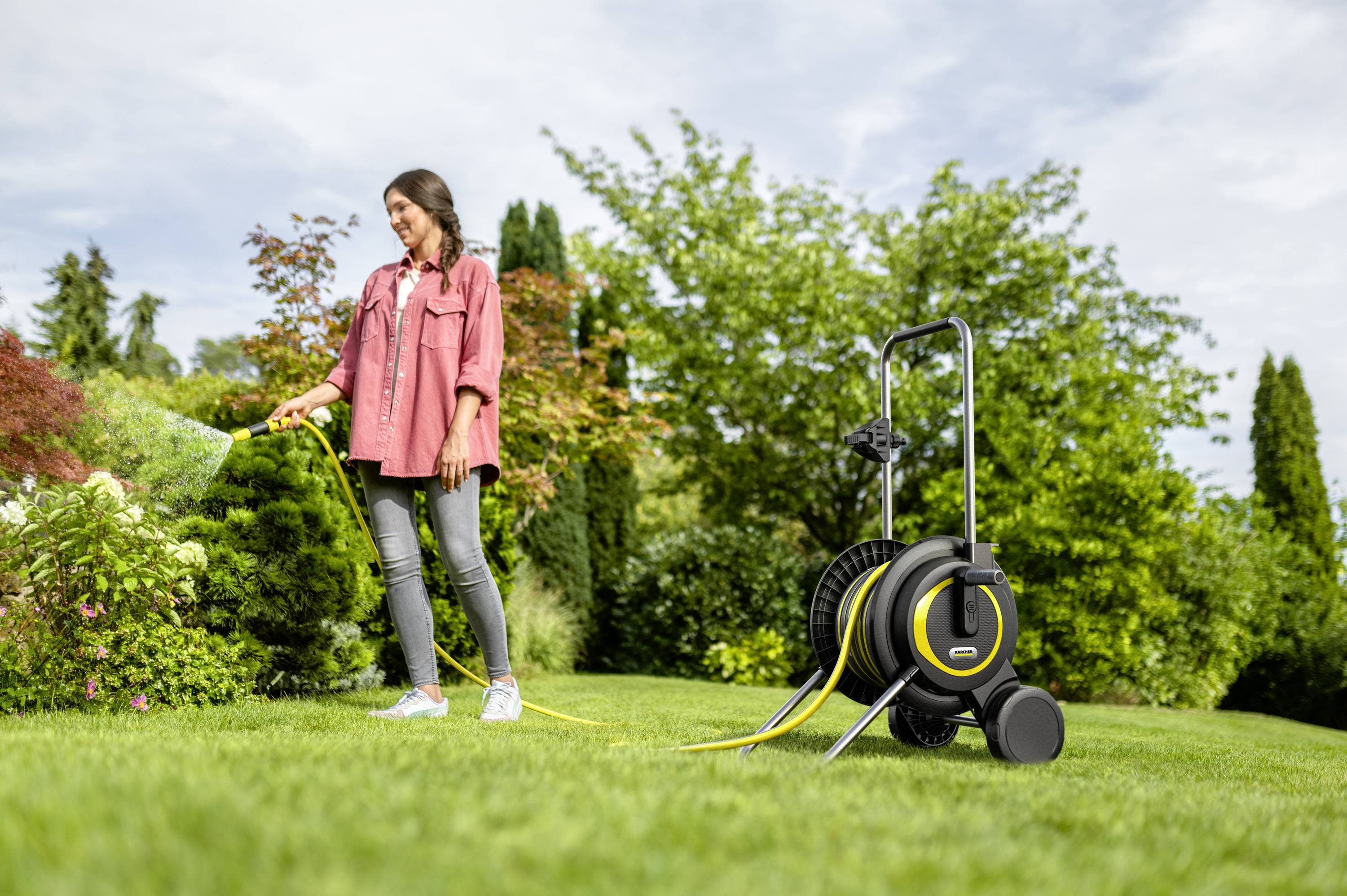 Eine Frau gießt einen Garten mit einem Schlauchwagen auf grünem Rasen, umgeben von Pflanzen und Bäumen an einem sonnigen Tag.