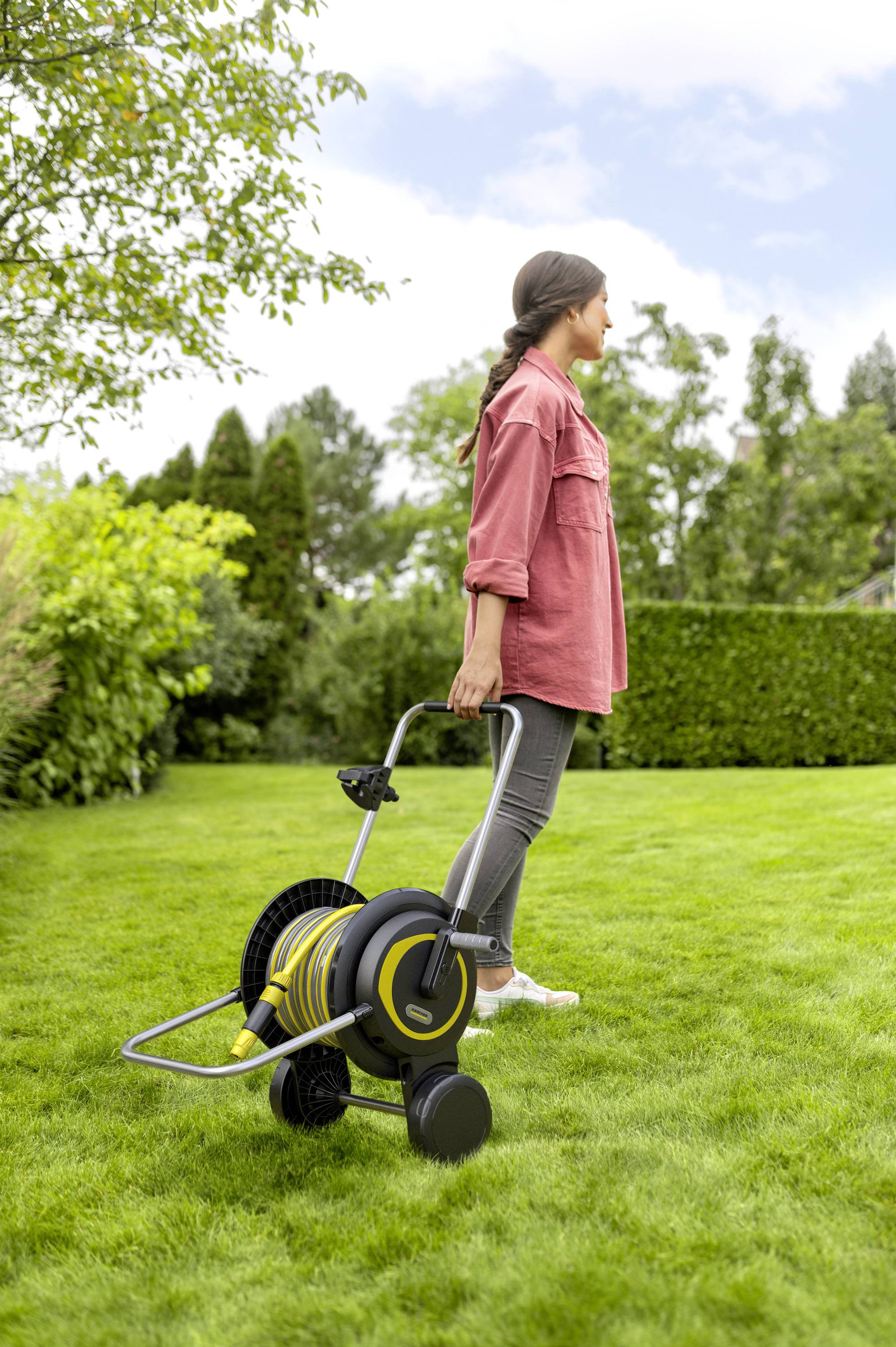Eine Person zieht einen Gartenschlauchwagen über einen grünen Rasen in einem Garten. Im Hintergrund sind Bäume und Büsche.