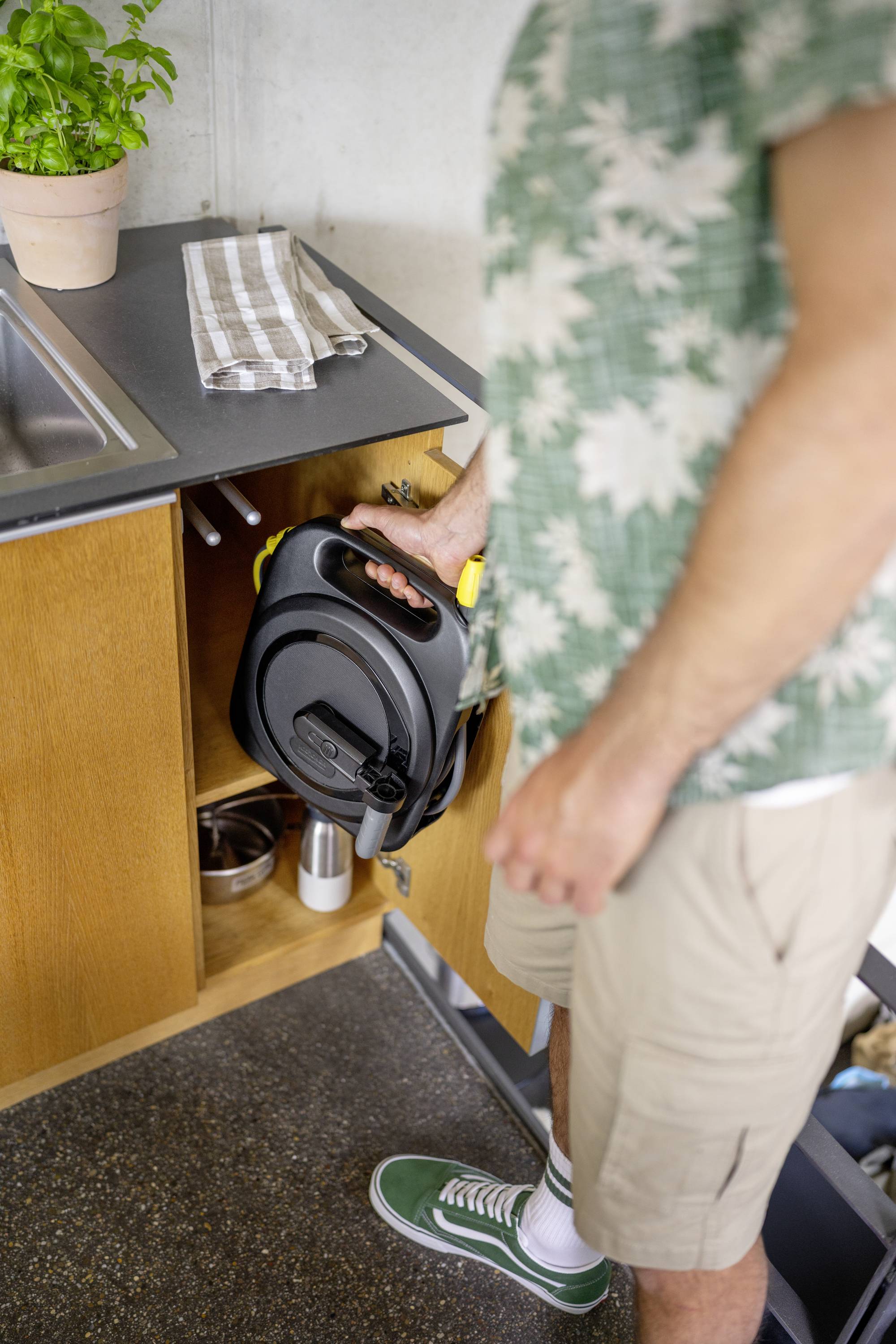 Eine Person in Sommerkleidung öffnet einen Unterschrank in einer Küche. Im Schrank liegt eine aufgerollte Verlängerungskabeltrommel.
