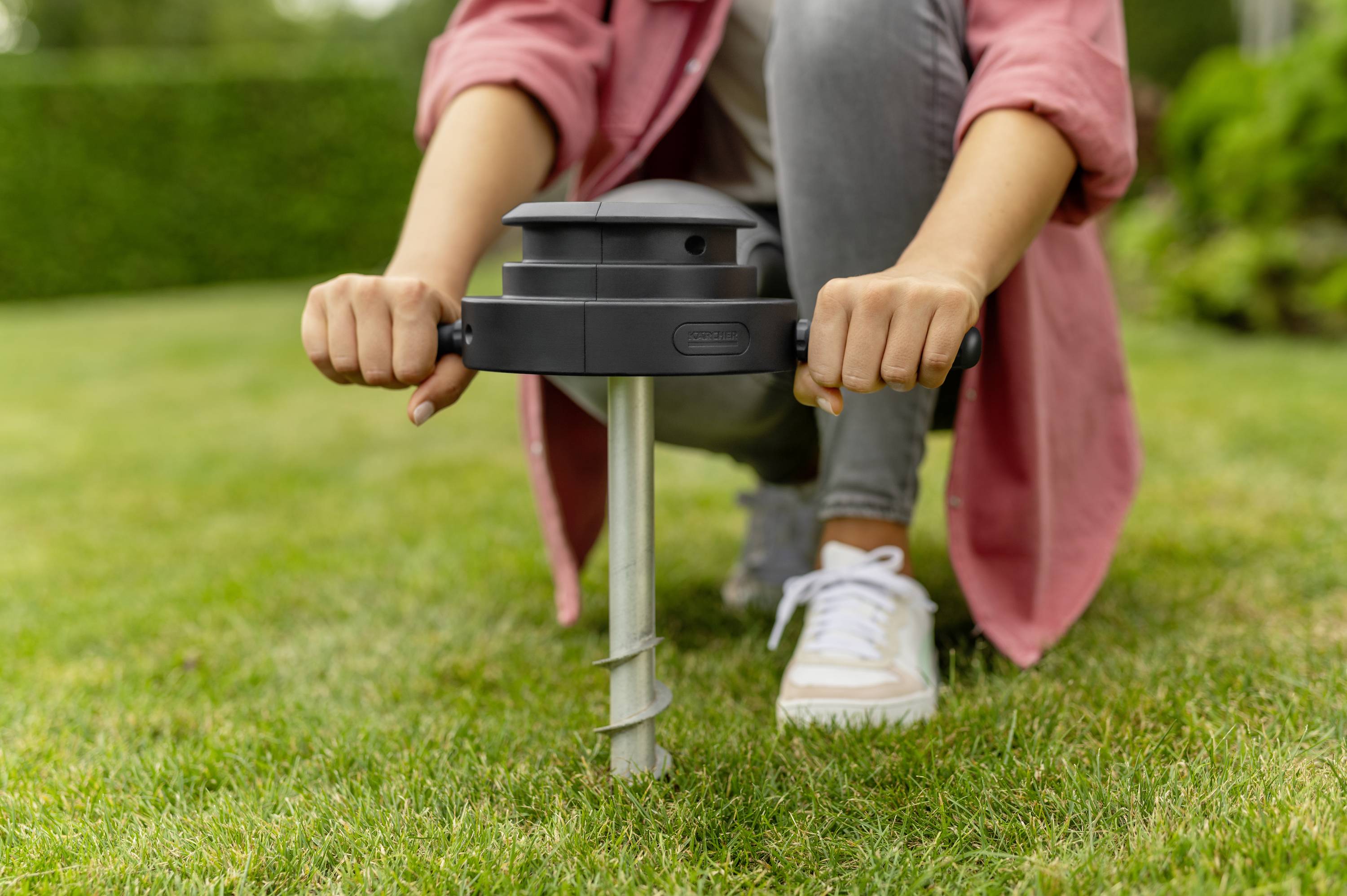 Person installiert ein Gerät im Rasen mit einem Handgriff und einer Schraube. Sie trägt Sneakers und eine rote Jacke. Im Hintergrund ist Garten zu sehen.