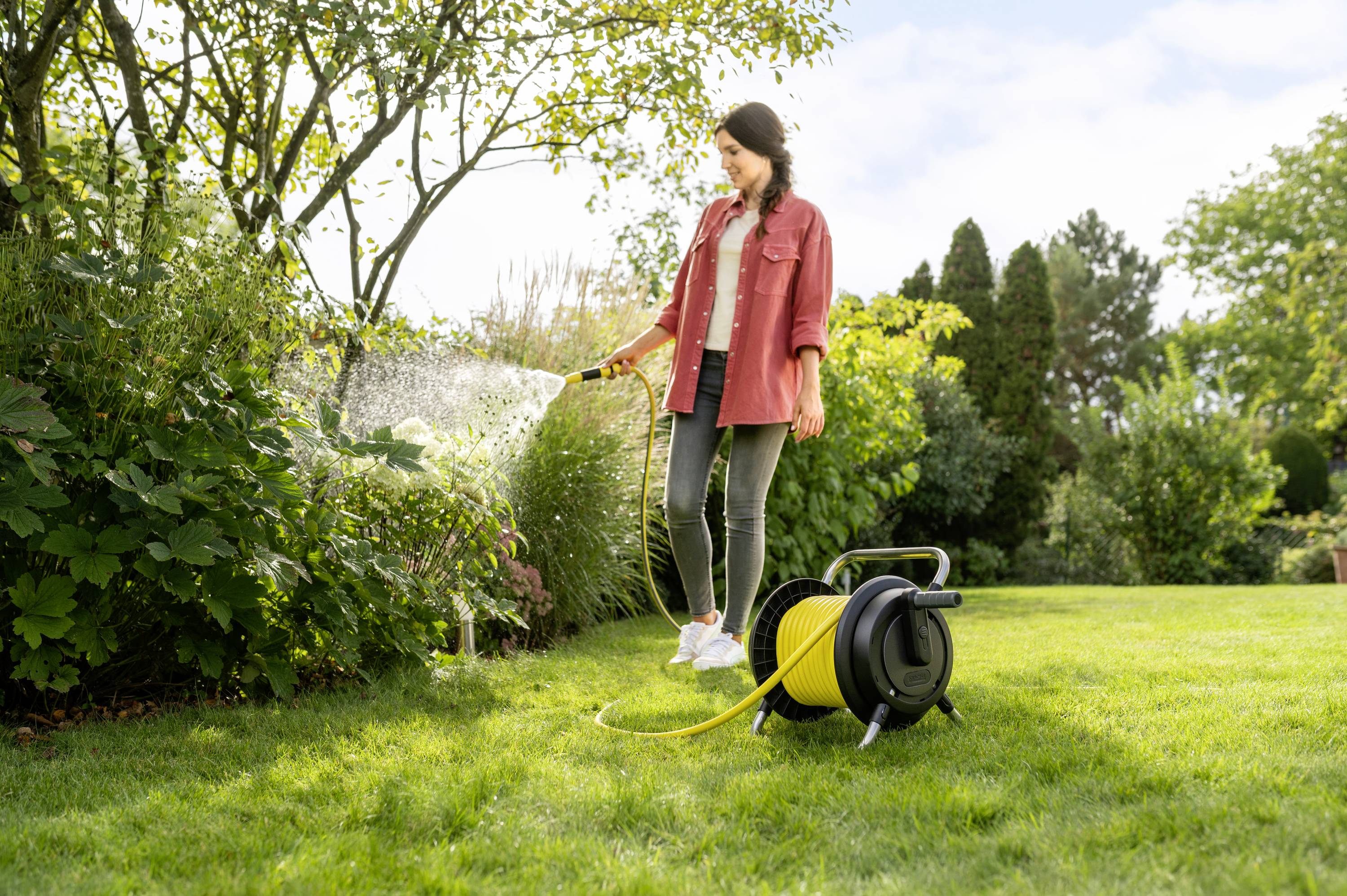 Eine Frau mit roter Jacke gießt Blumen im Garten mit einem Wasserschlauch. Ein Schlauchaufroller liegt neben ihr auf dem Rasen.