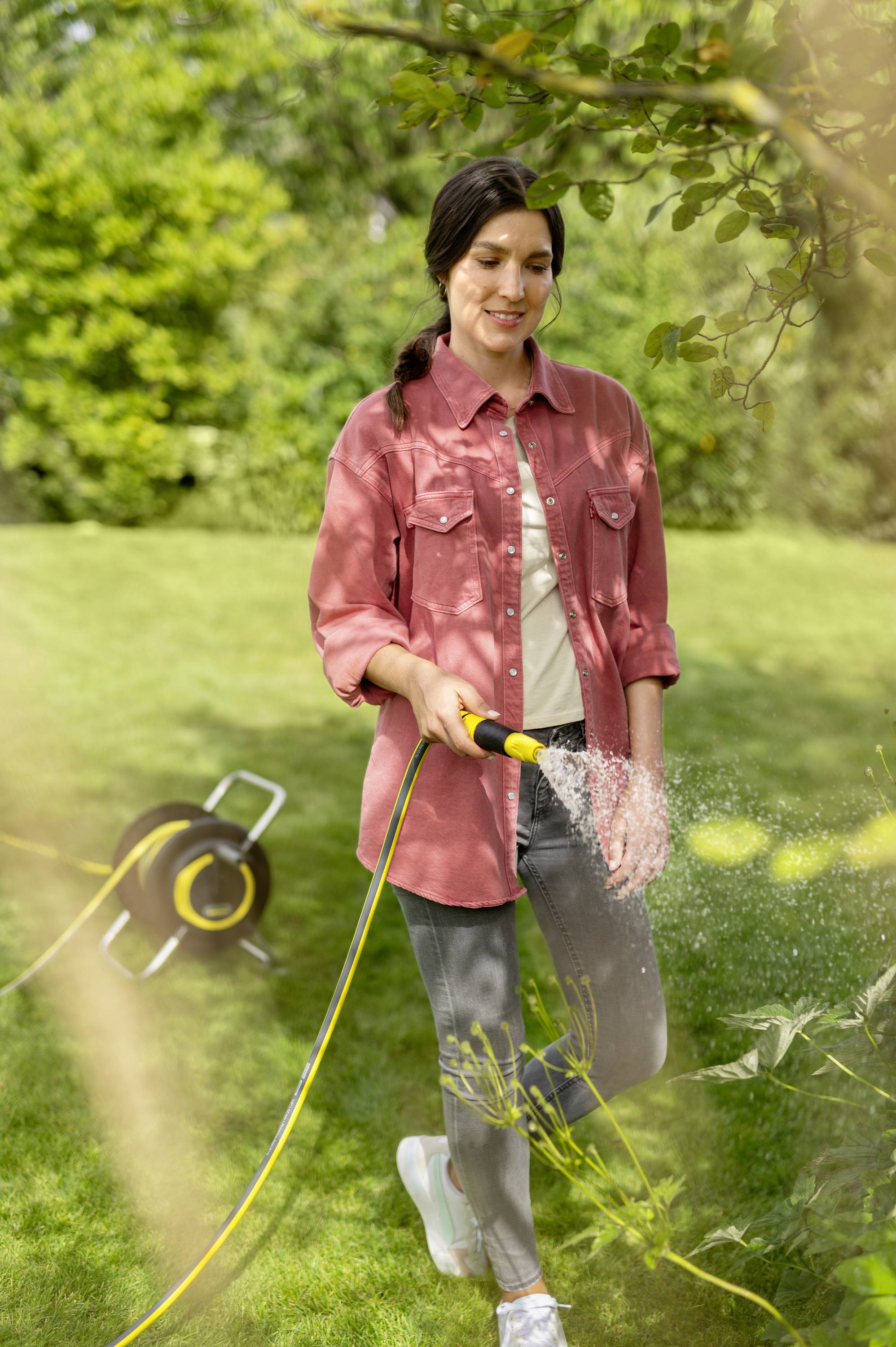 Eine Person in Freizeitkleidung gießt mit einem Gartenschlauch Pflanzen in einem grünen Garten. Im Hintergrund ist eine Schlauchtrommel sichtbar.