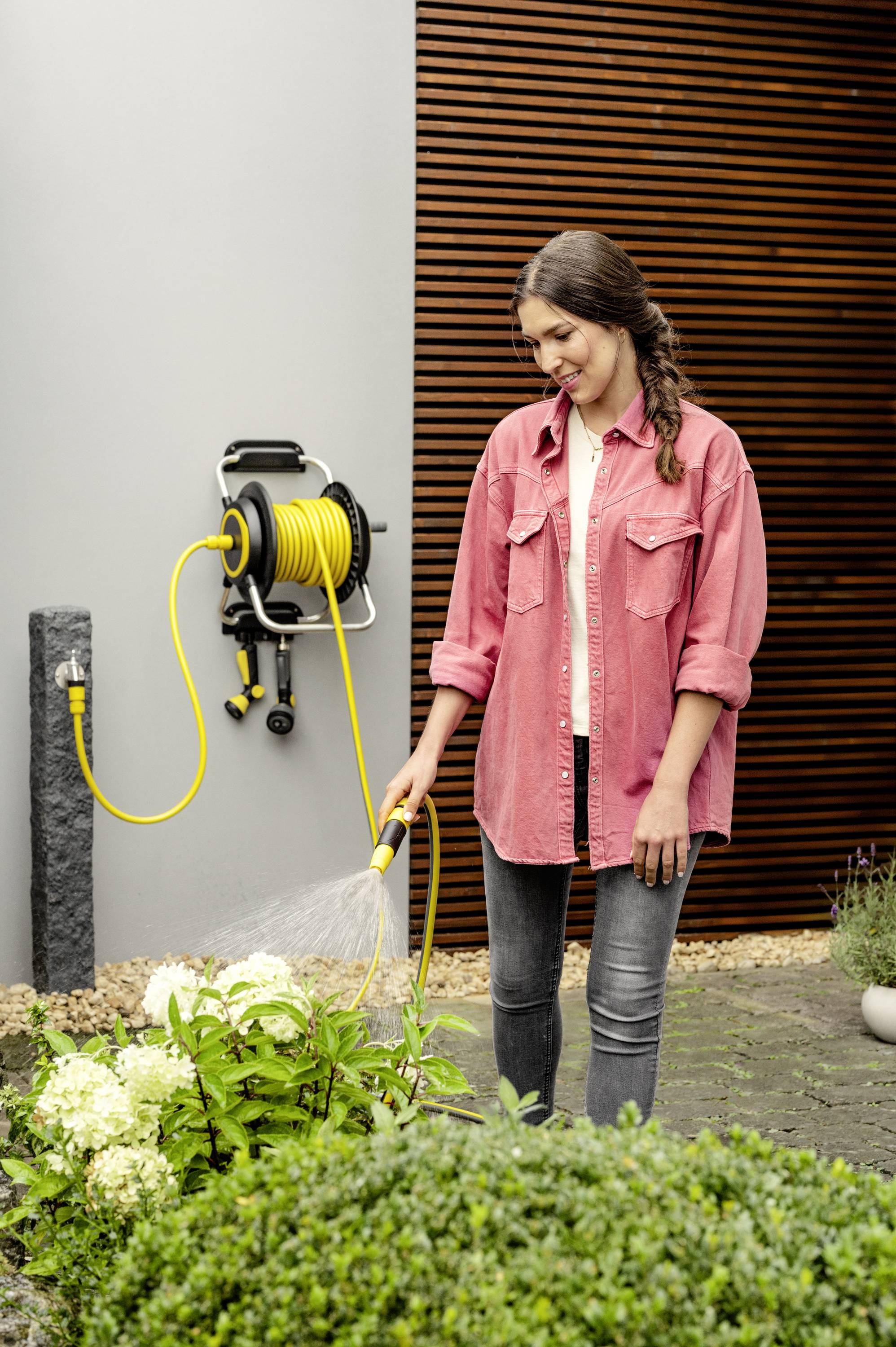 Eine Frau in einer rosafarbenen Jacke gießt Pflanzen mit einem gelben Gartenschlauch. Im Hintergrund eine Wand mit Holzpaneelen.
