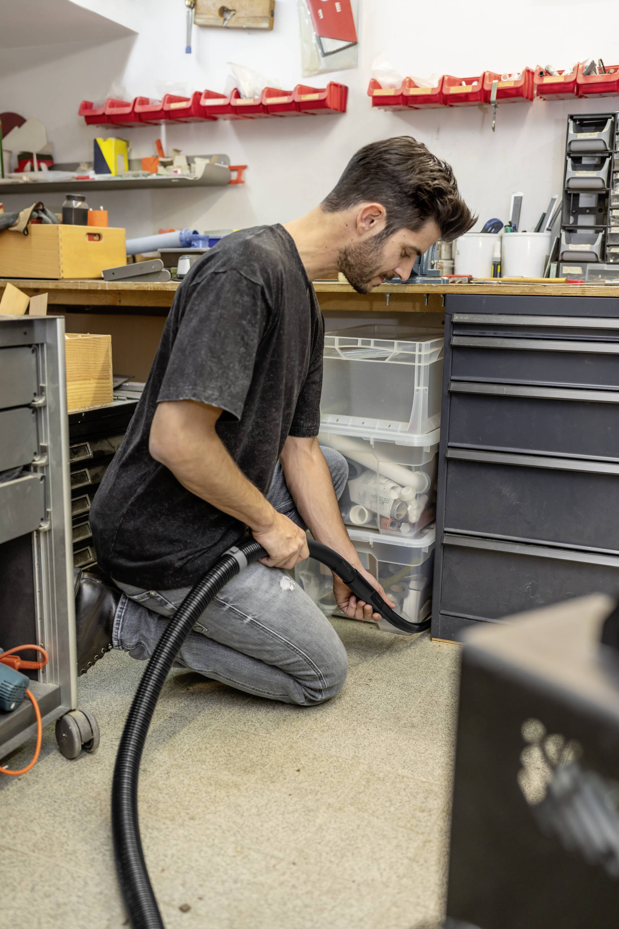Junger Mann kniet auf einem Werkstattboden und saugt mit einem Staubsauger. Im Hintergrund sind Werkzeuge und Regale zu sehen.