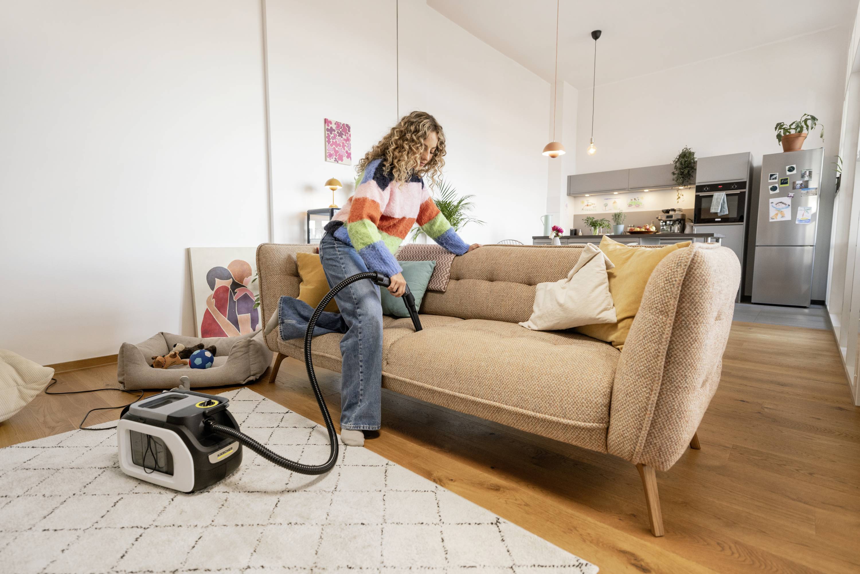Eine Person saugt mit einem Dampfreiniger ein Sofa in einem modernen Wohnzimmer. Im Hintergrund ist eine Küche zu sehen.