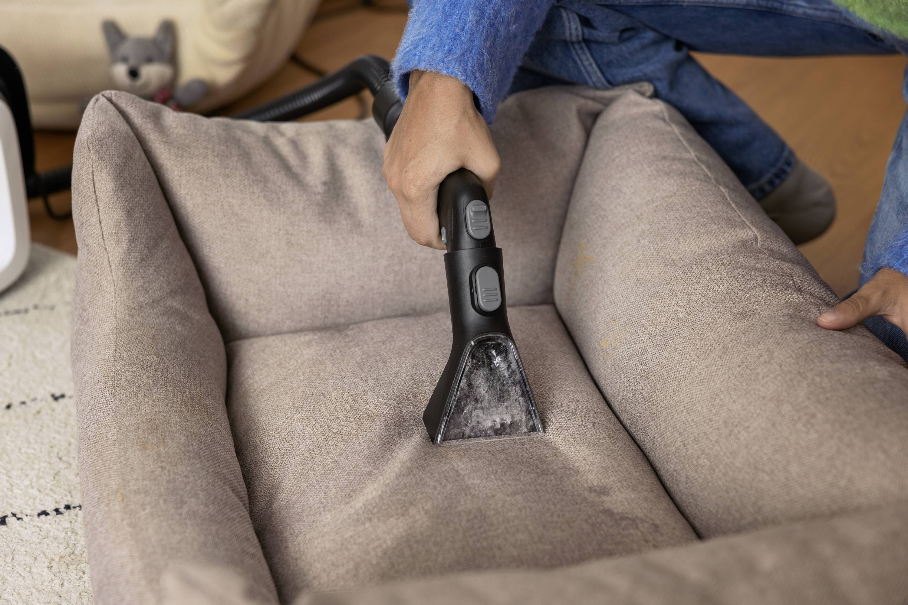 Eine Person reinigt ein braunes Hundebett mit einem Handstaubsauger. Im Hintergrund ist ein Hundespielzeug zu sehen.