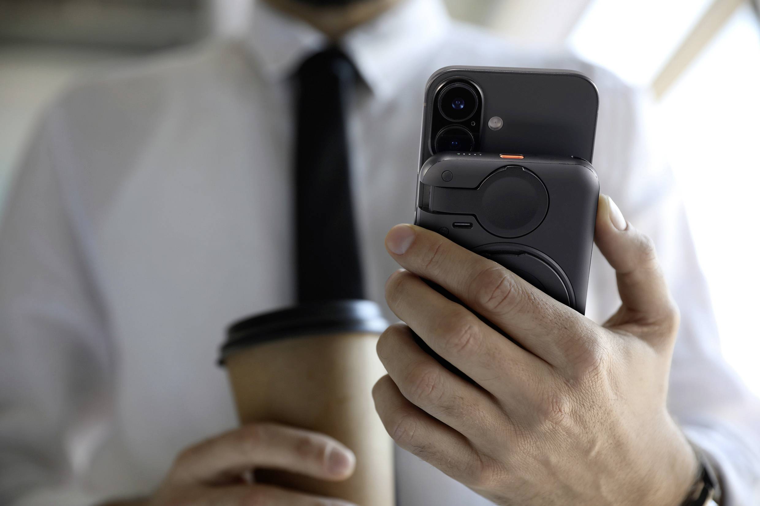 Eine Person hält mit der rechten Hand ein Smartphone und mit der linken Hand einen Pappbecher, trägt ein weißes Hemd und eine schwarze Krawatte.