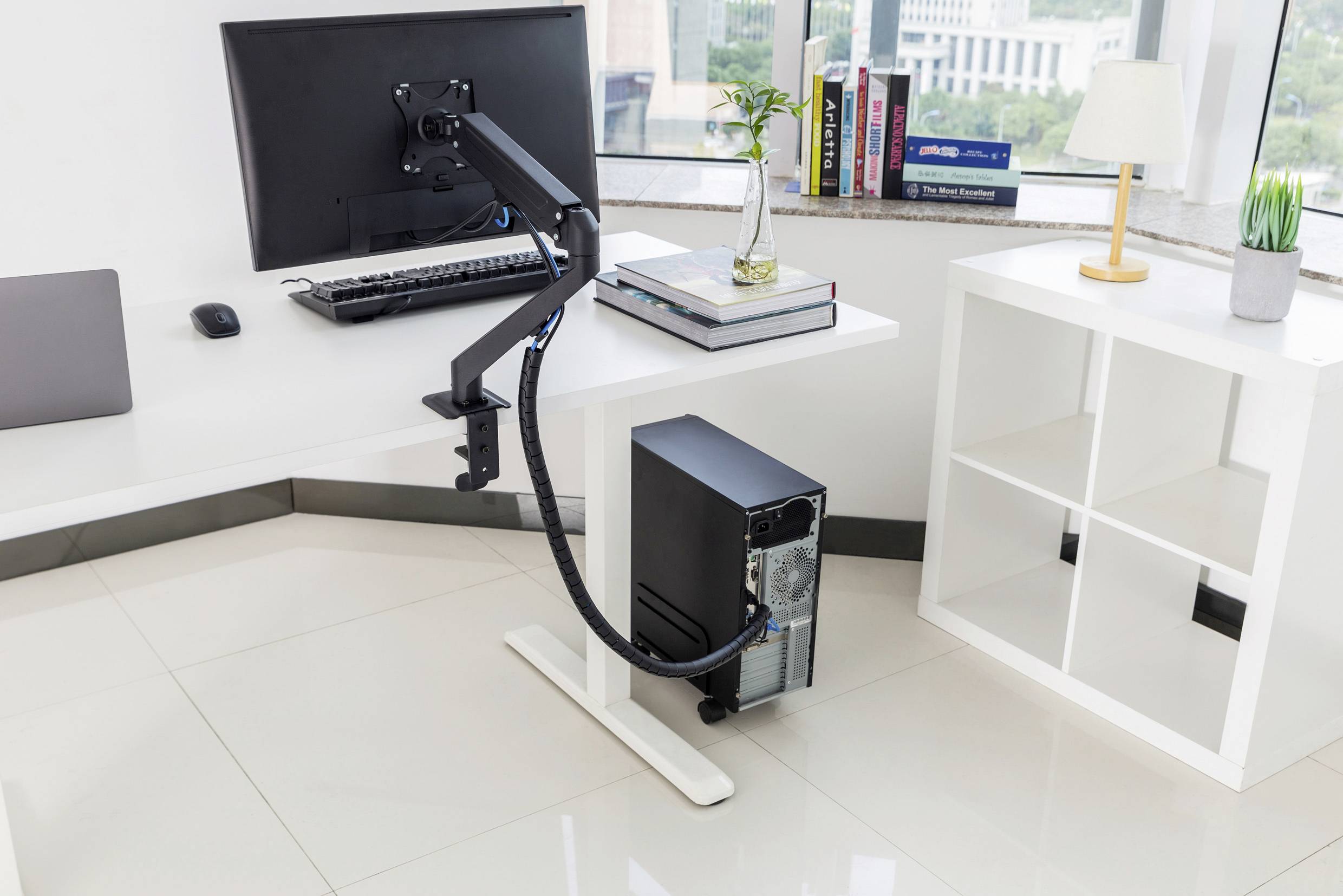 Büro mit weißem Tisch, darauf Monitor mit Schwenkarm. Darunter schwarzer Computer. Fenster mit Stadtblick im Hintergrund.