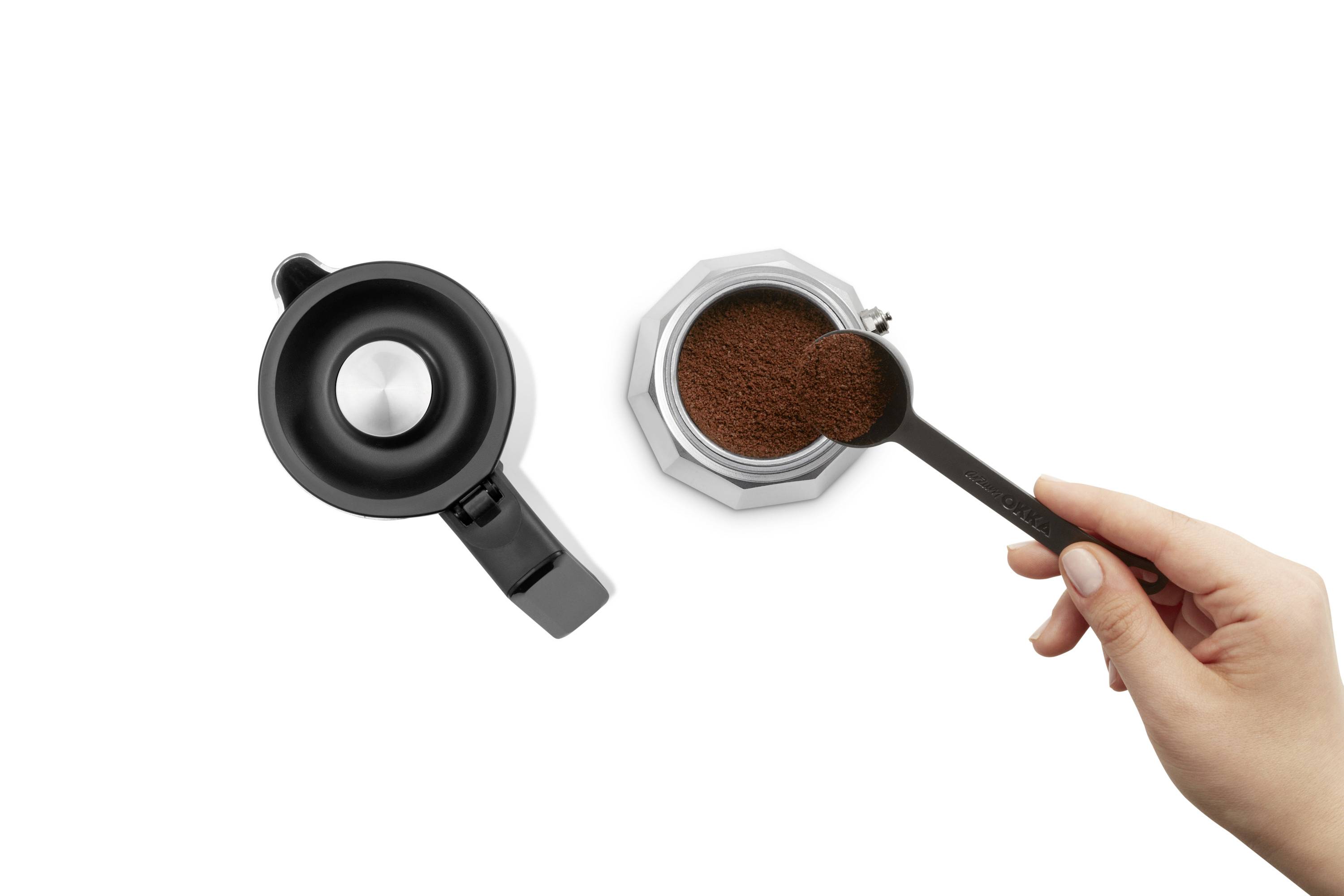 Eine Hand füllt gemahlenen Kaffee mit einem Löffel in eine Mokkakanne neben einem Deckel.