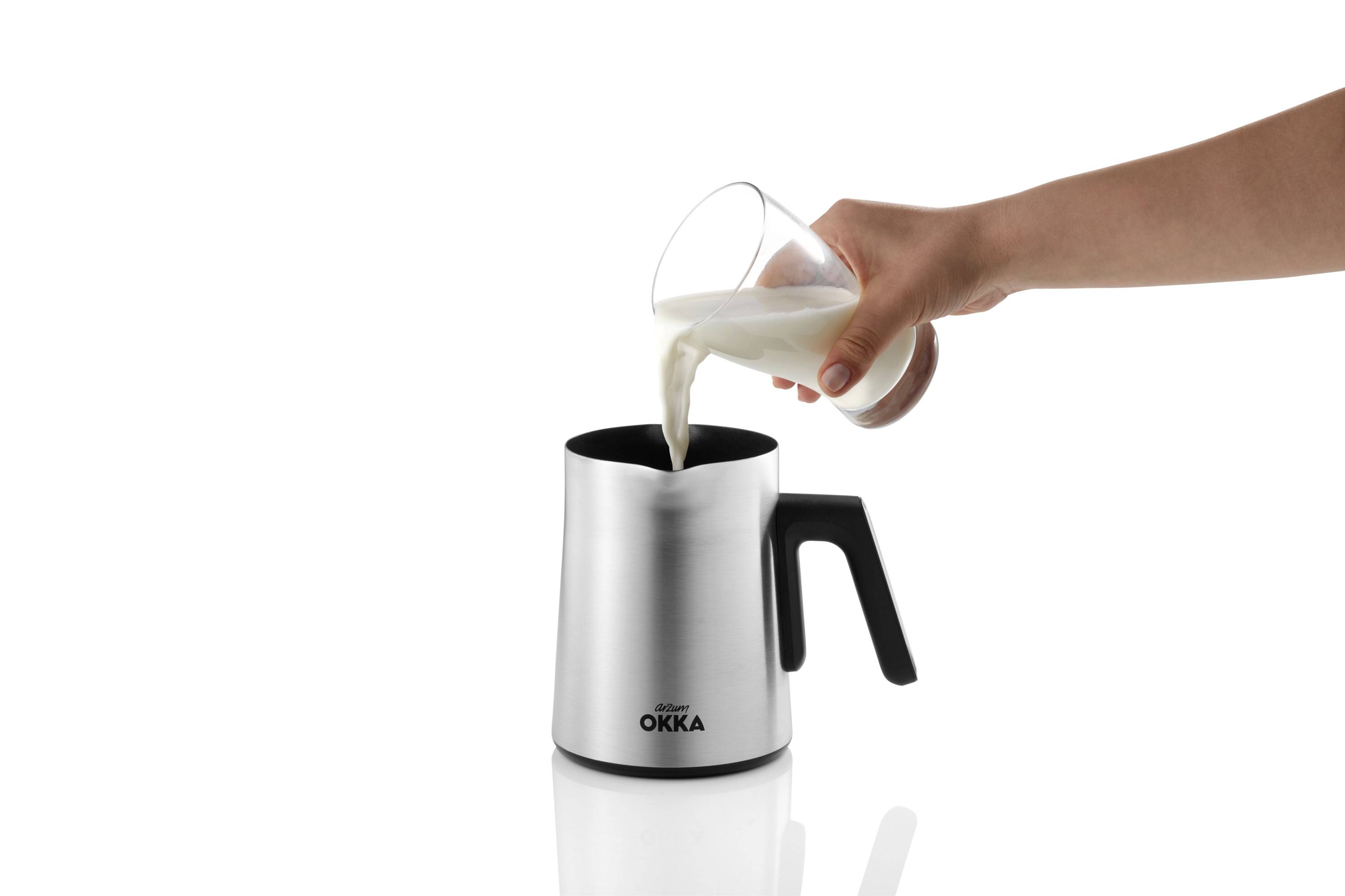 Eine Hand gießt Milch aus einem Glas in einen silbernen Milchkännchen mit schwarzem Griff.