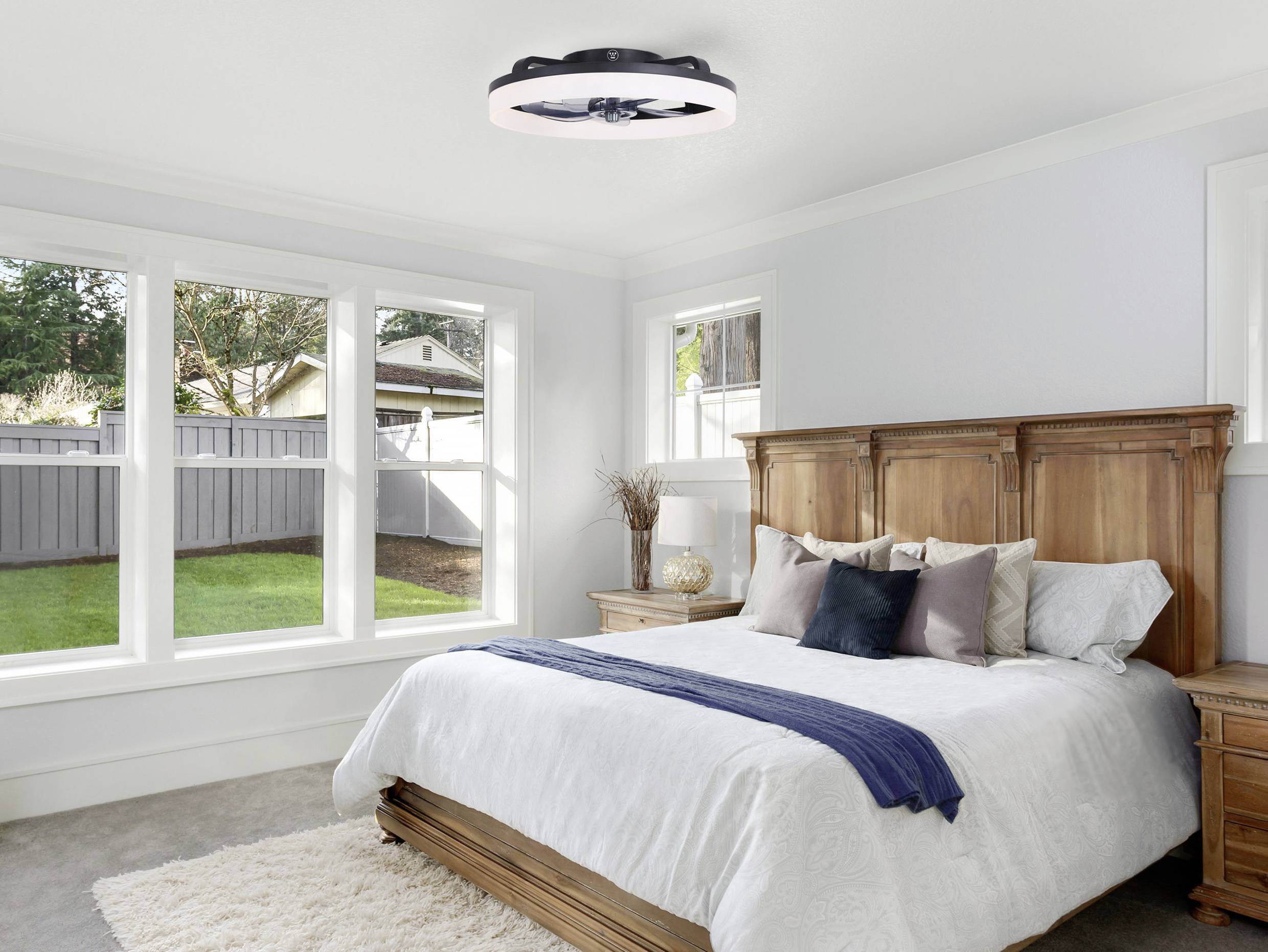 Schlafzimmer mit einem großen Fenster, Holzbett und einem modernen Deckenventilator. Das Zimmer hat eine ruhige, helle Atmosphäre.