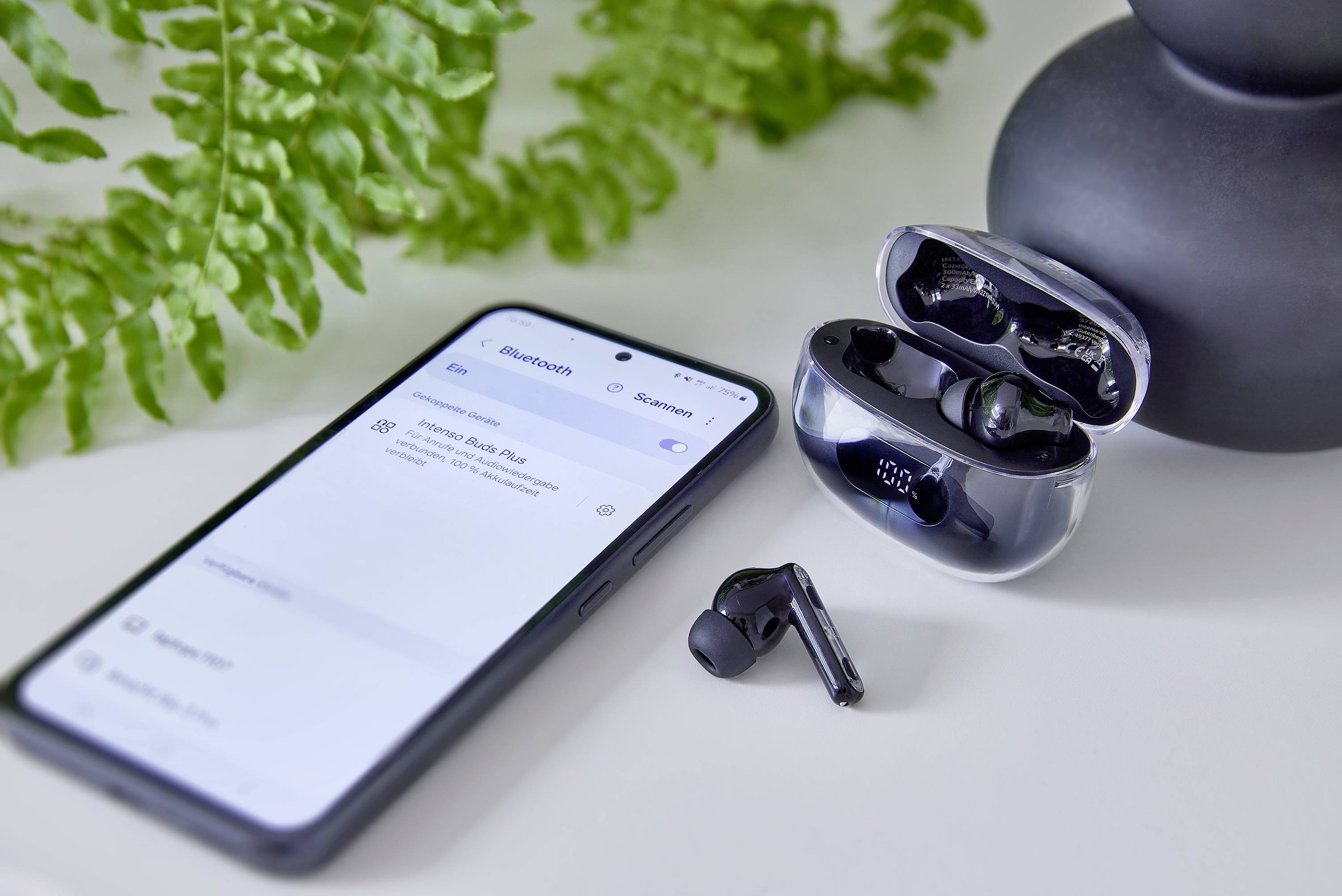Ein Smartphone zeigt Bluetooth-Einstellungen neben kabellosen In-Ear-Kopfhörern in einer offenen Ladebox. Im Hintergrund ist eine Pflanze.