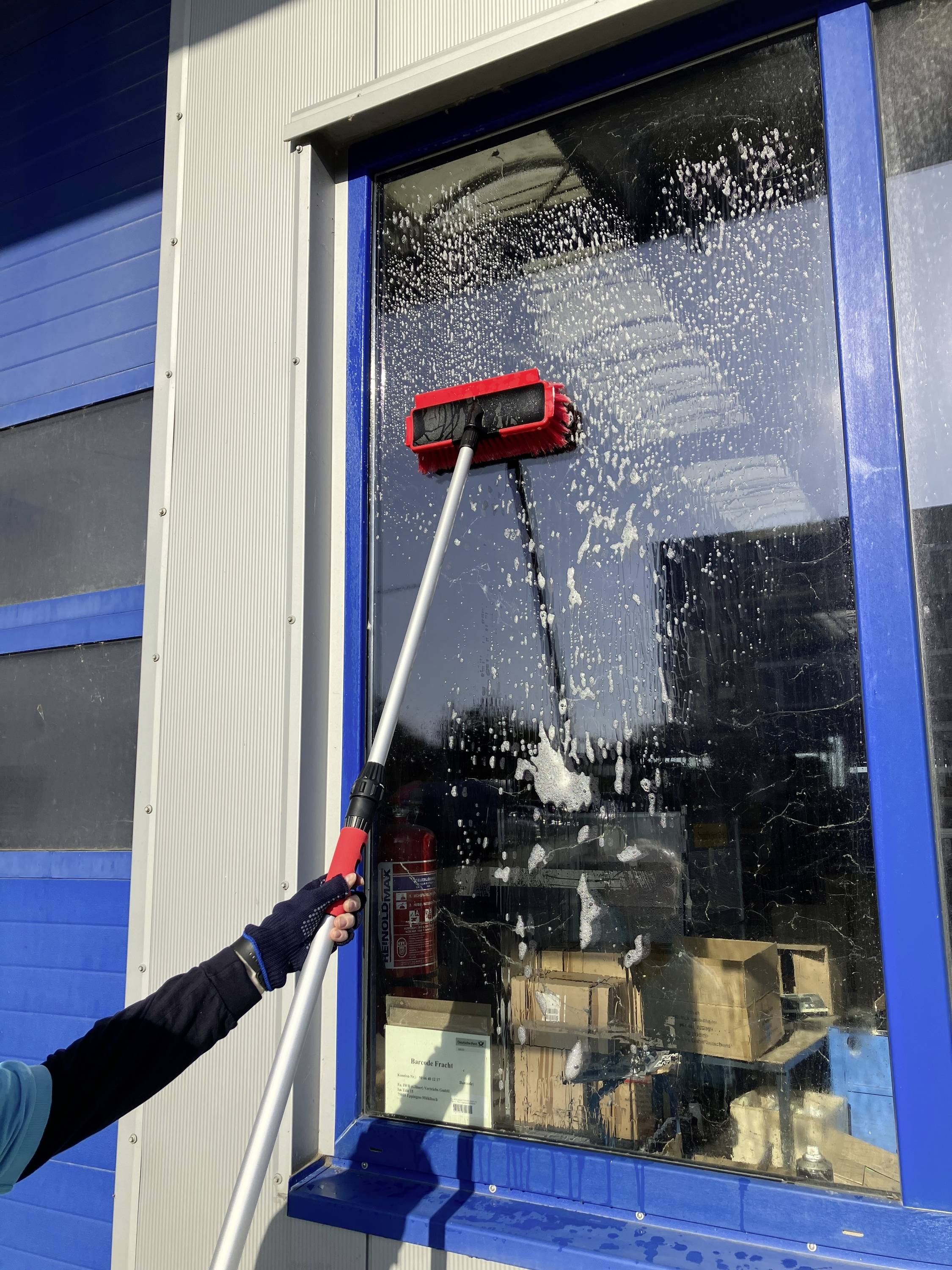 Eine Person reinigt bei Sonnenschein ein großes Fenster mit einer langen Bürste und Seifenwasser.