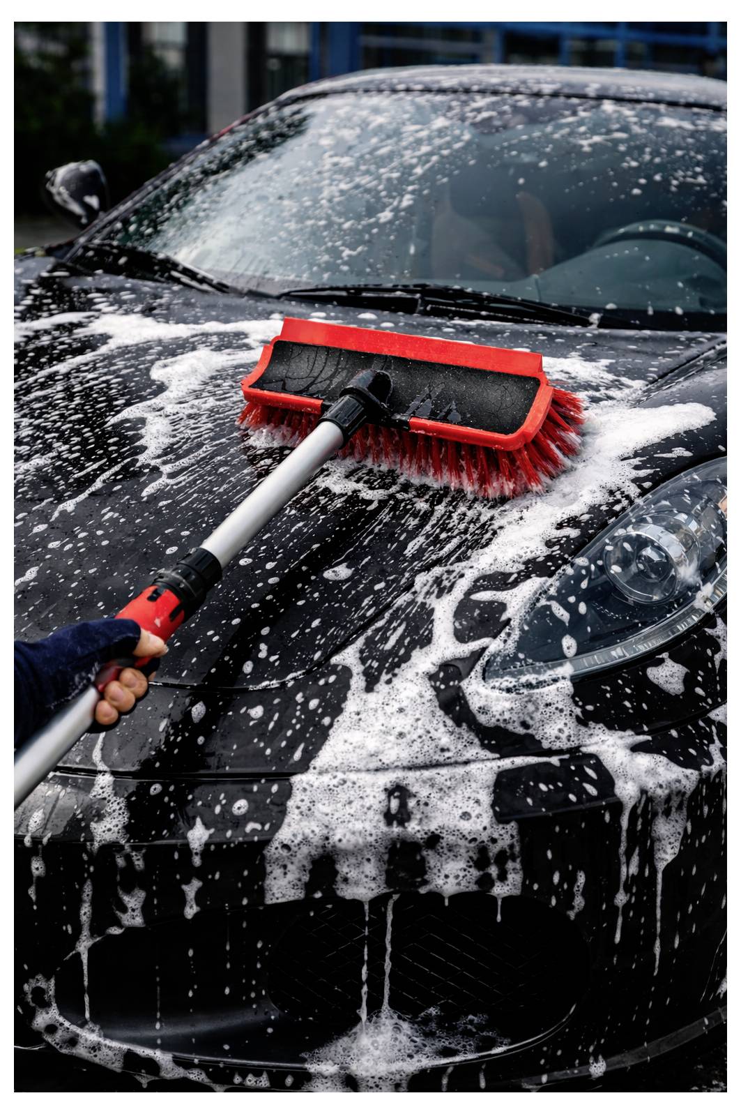 Eine Person, die ein schwarzes Auto mit einer roten Bürste wäscht, bedeckt mit Seifenschaum.