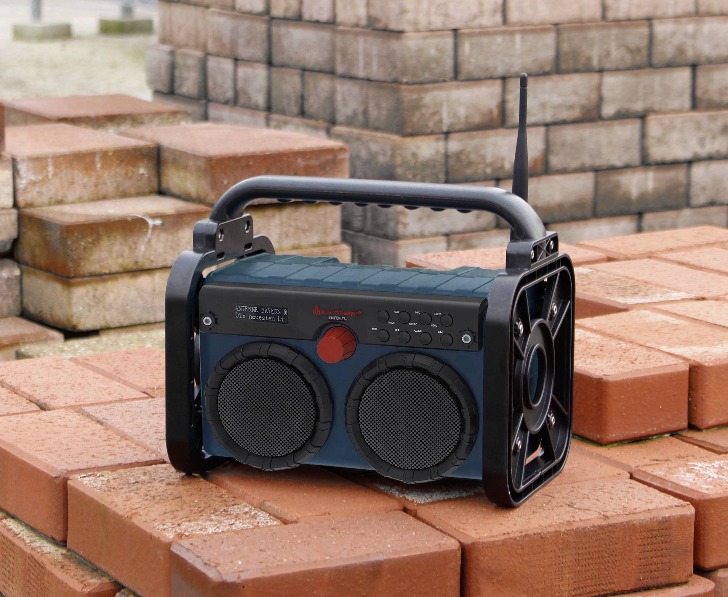Ein tragbares Baustellenradio mit zwei Lautsprechern steht auf gestapelten Ziegeln. Es hat einen Griff und eine Antenne.
