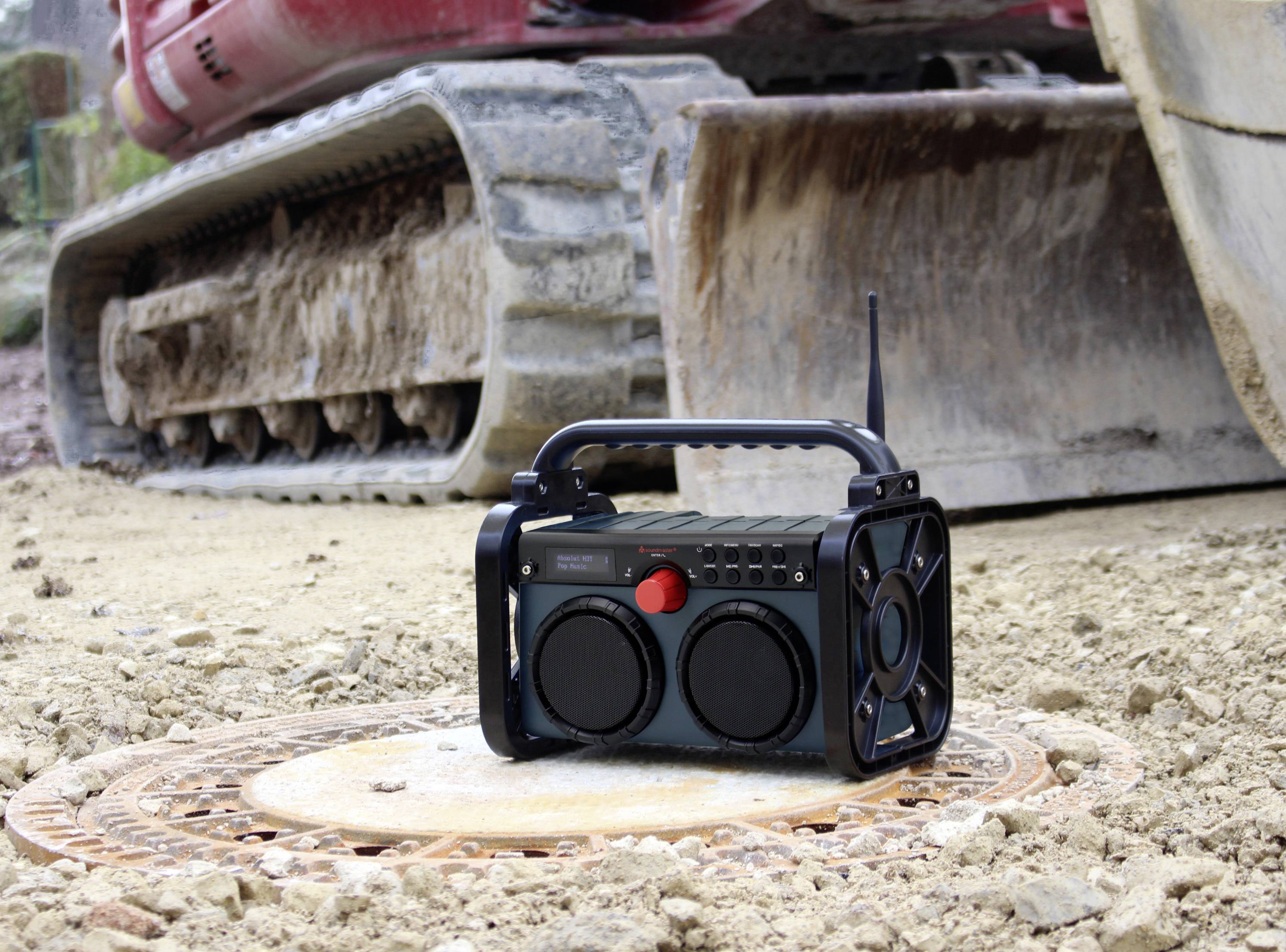 Ein tragbares Radio steht auf einem Kanaldeckel auf einer Baustelle, im Hintergrund ein Baggerfahrzeug.