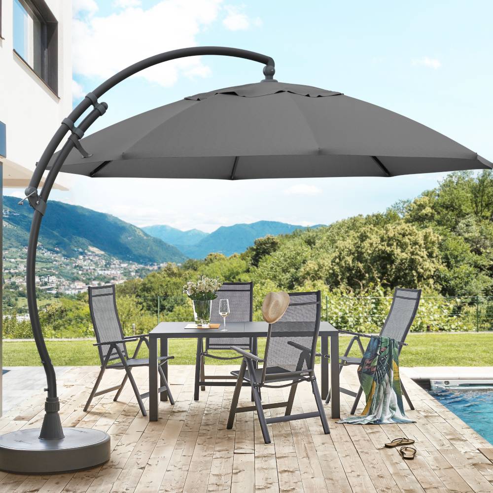 Runder Gartentisch mit vier Stühlen und großem Sonnenschirm auf einer Terrasse. Im Hintergrund Berge und Bäume, blaue Himmel.