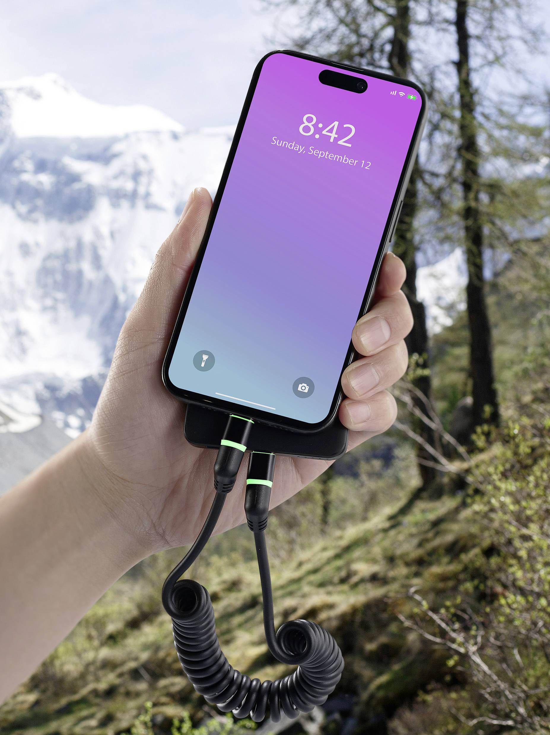 Eine Hand hält ein Smartphone mit lila-blauem Bildschirm und zeigt '8:42 Sunday, September 12'. Im Hintergrund sind Berge und Bäume.