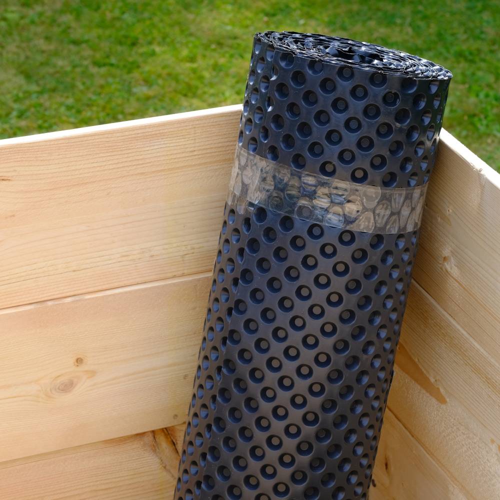 Eine schwarze Noppenbahn steht aufrecht in einer Holzkiste im Freien. Die Bahn ist zum Schutz oder zur Entwässerung geeignet.