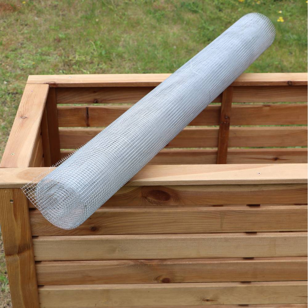 Eine Rolle Drahtgeflecht liegt auf einem hölzernen Komposter im Freien. Grünflächen im Hintergrund.