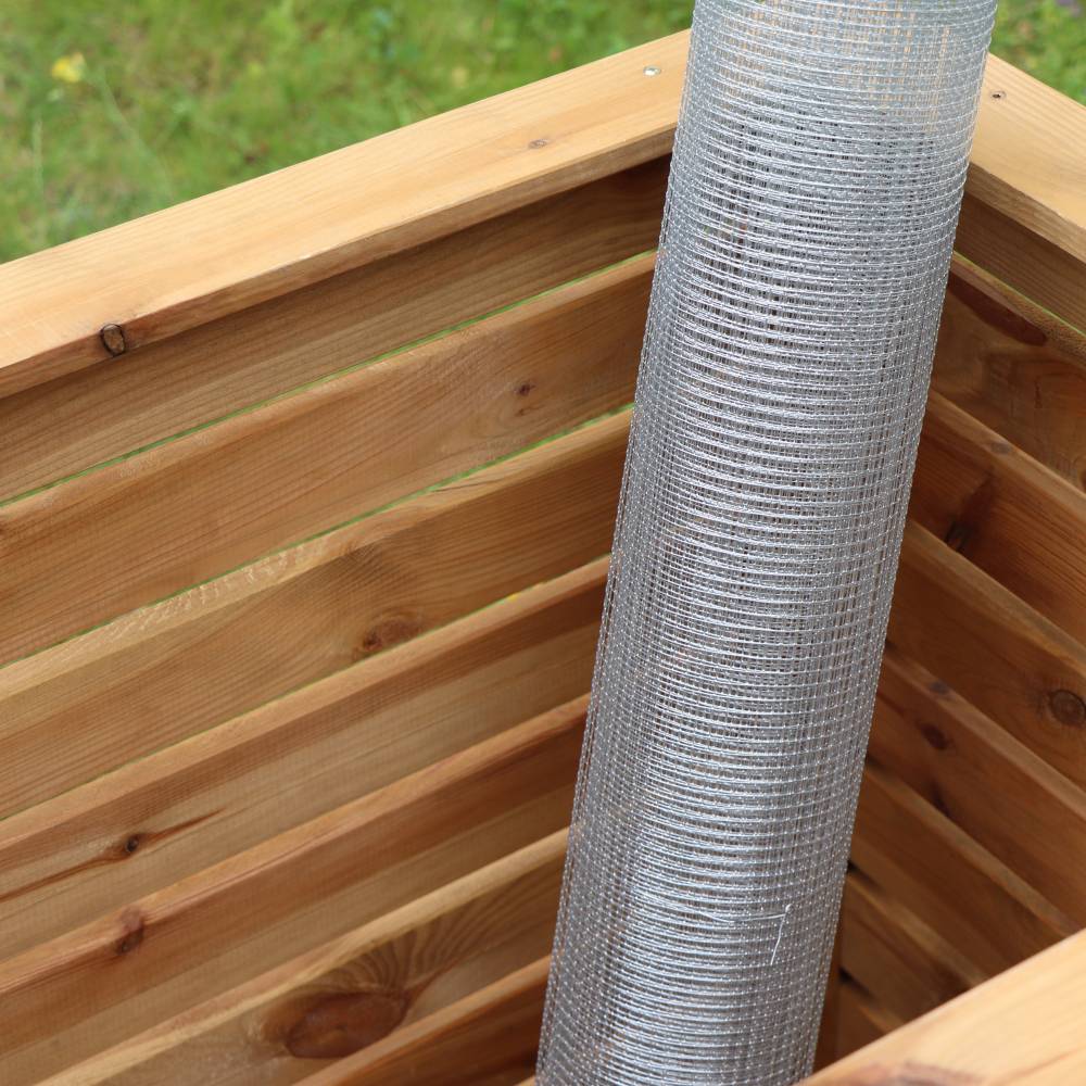 Eine Rolle Drahtgeflecht steht in einer hölzernen Kiste. Die Kiste hat Holzbretter mit horizontalen Streifen und befindet sich im Freien auf Wiese.