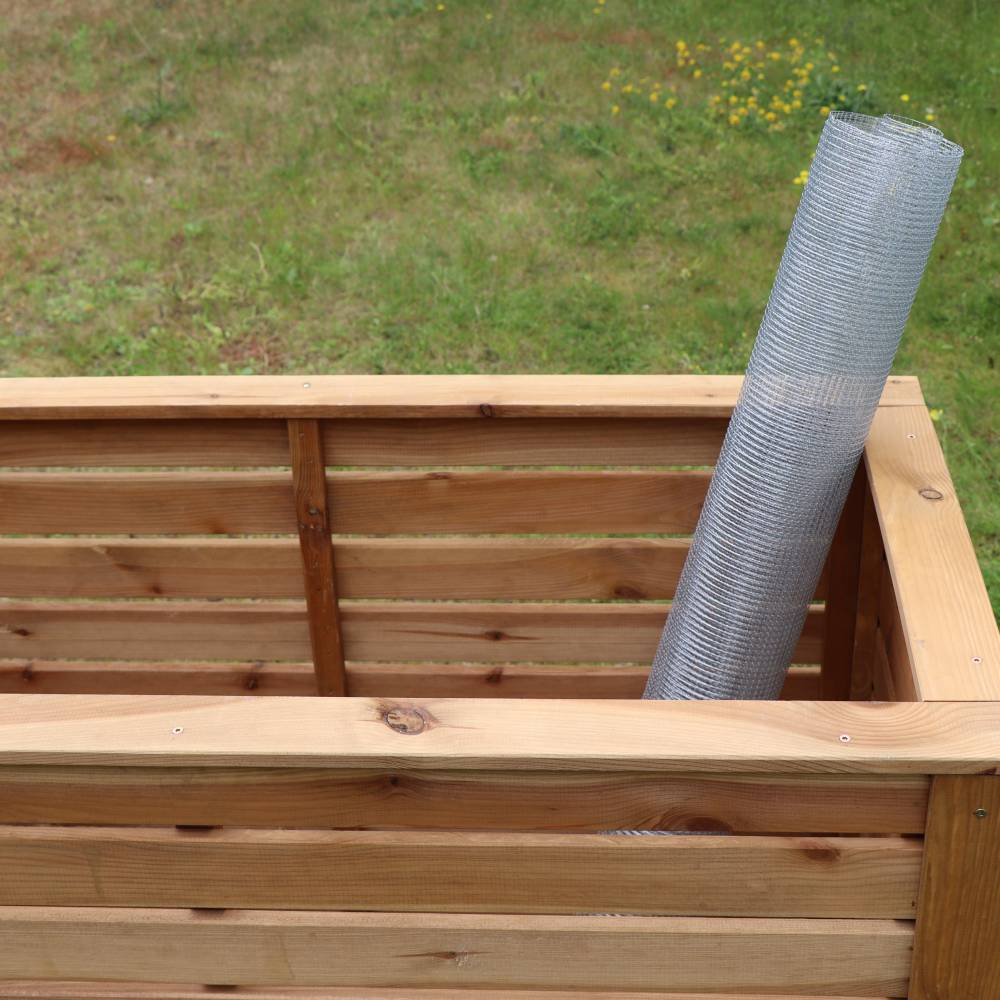 Ein Holzkomposter im Garten mit einer Rolle Drahtgeflecht zur Stabilisierung. Hintergrund zeigt grünes Gras und gelbe Blumen.