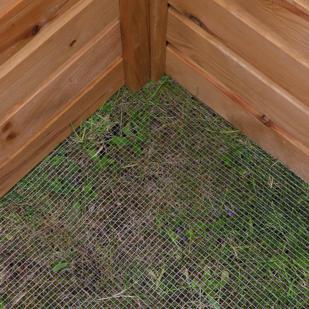 Sicht von oben auf die Ecke eines Holzkomposts mit Drahtgeflecht am Boden und Gras darunter.