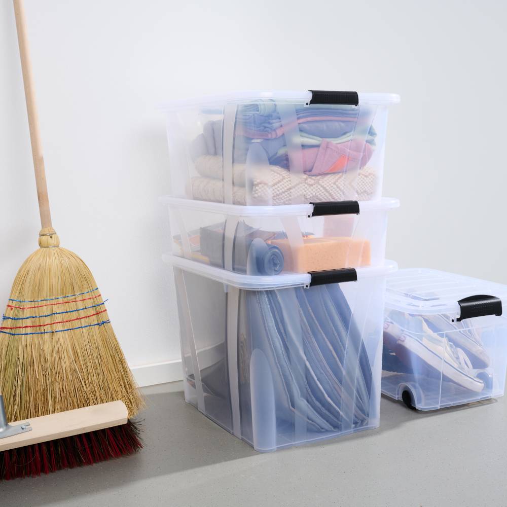 Ein Besen lehnt an der Wand neben drei durchsichtigen, gestapelten Boxen voller Kleidung und Bettwäsche auf einem grauen Boden.