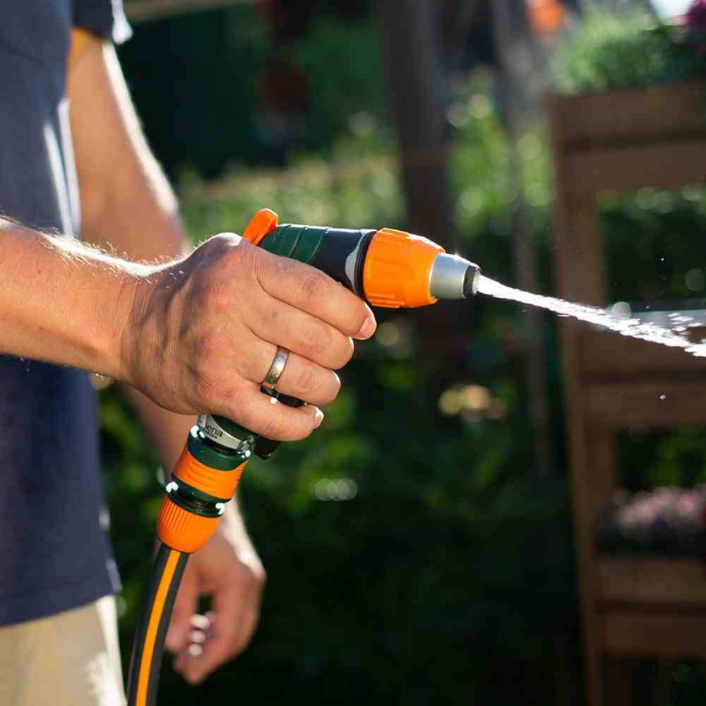 Eine Person gießt mit einem Gartenschlauch Pflanzen im Sonnenlicht. Der Fokus liegt auf dem Wasserstrahl und der Hand, die den Schlauch hält.