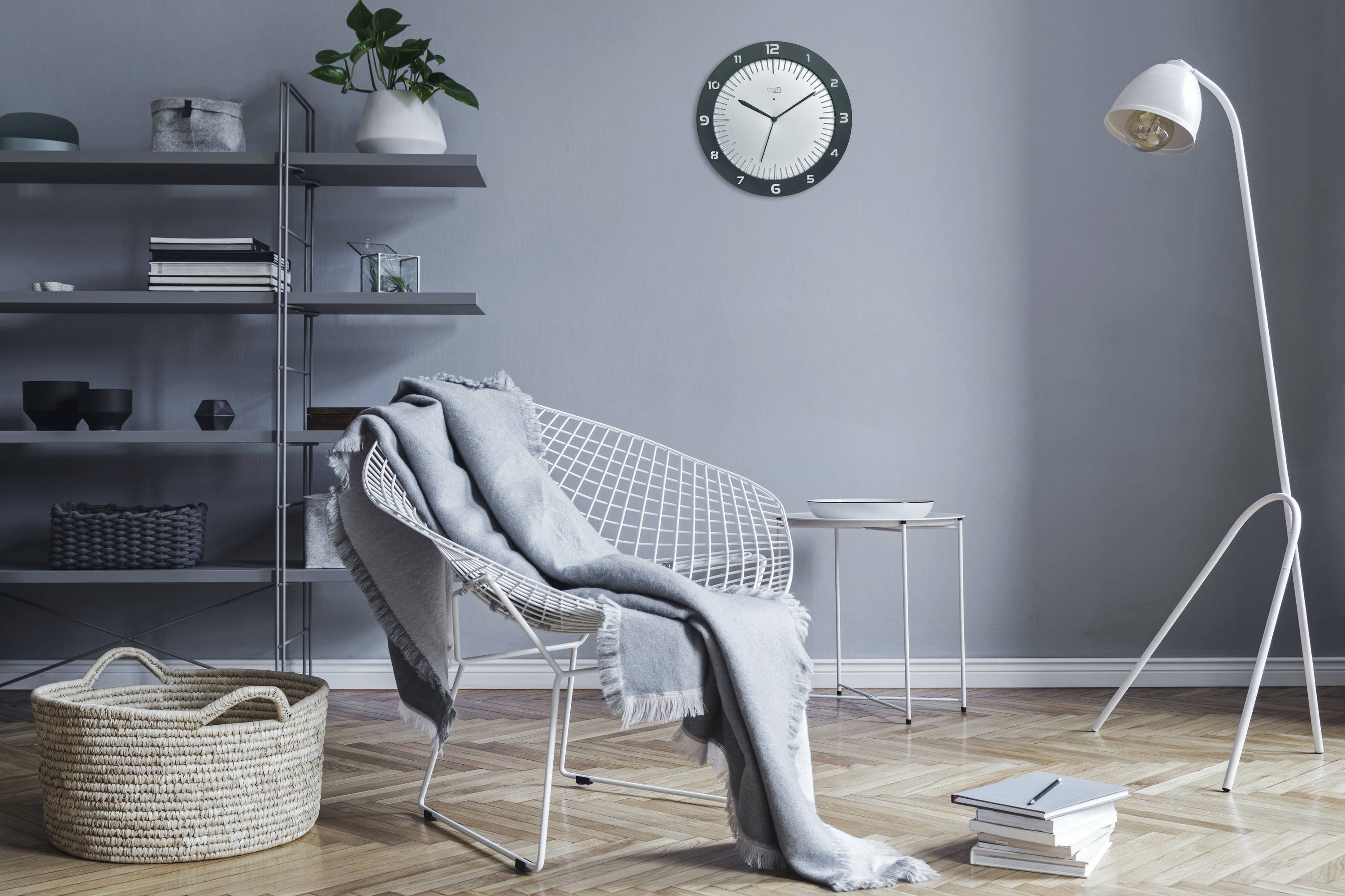 Wohnzimmer mit modernem Sessel, grauem Überwurf und Lampe. An der Wand hängt eine Uhr. Regal mit Pflanzen und Dekorationen im Hintergrund.