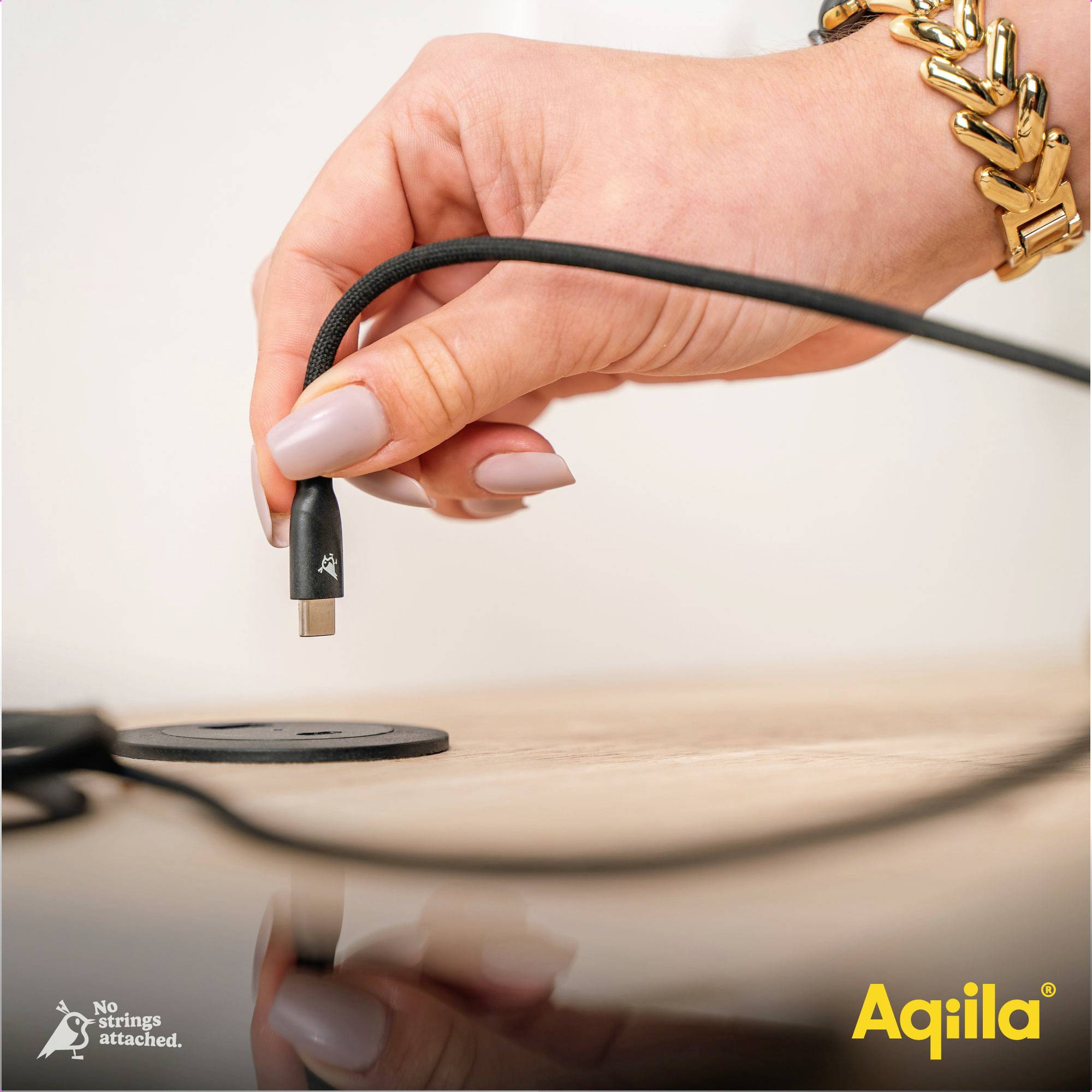 Eine Hand hält ein USB-Kabel über einem Tisch. Unten links steht 'No strings attached'. Unten rechts das Logo 'Aqilla'.