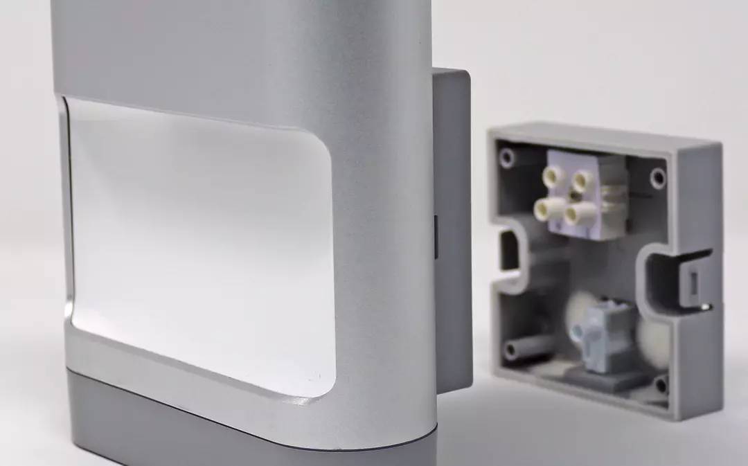 Ein modernes, rechteckiges Gebrauchsobjekt aus Metall mit abgerundeten Kanten, links im Vordergrund. Ein technisches Teil ist im Hintergrund.