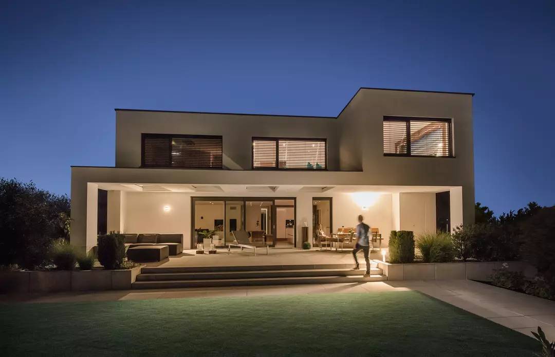 Ein modernes zweistöckiges Haus bei Nacht, mit beleuchteter Terrasse und großen Fenstern. Eine Person geht über den Rasen.