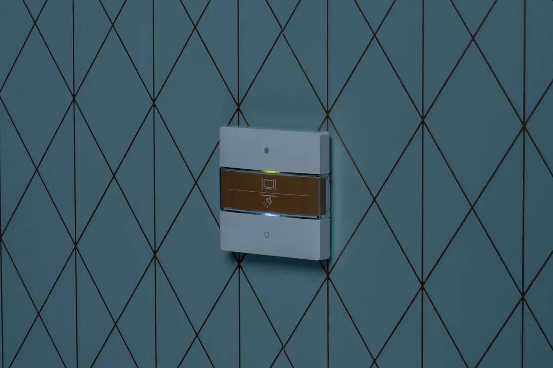 Ein weißer Lichtschalter mit einem goldenen Streifen in der Mitte befindet sich an einer blauen Kachelwand mit Rautenmuster.