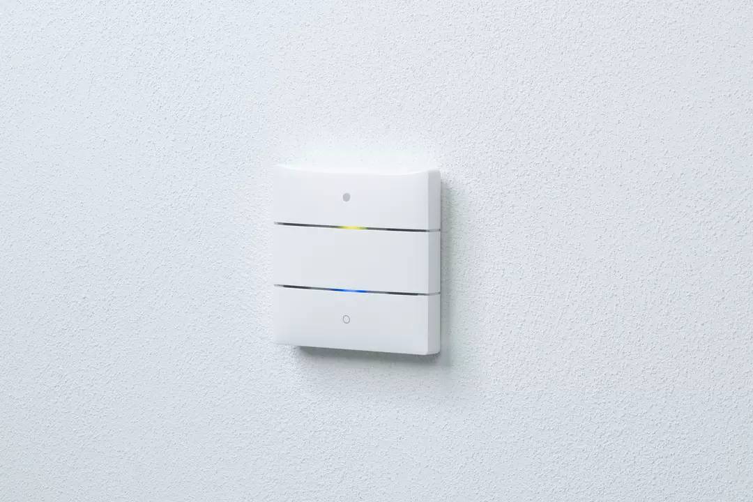 Ein weißer, rechteckiger Wandschalter mit drei sichtbaren horizontalen Segmenten und einem kleinen Lämpchen, an einer grauen Wand montiert.