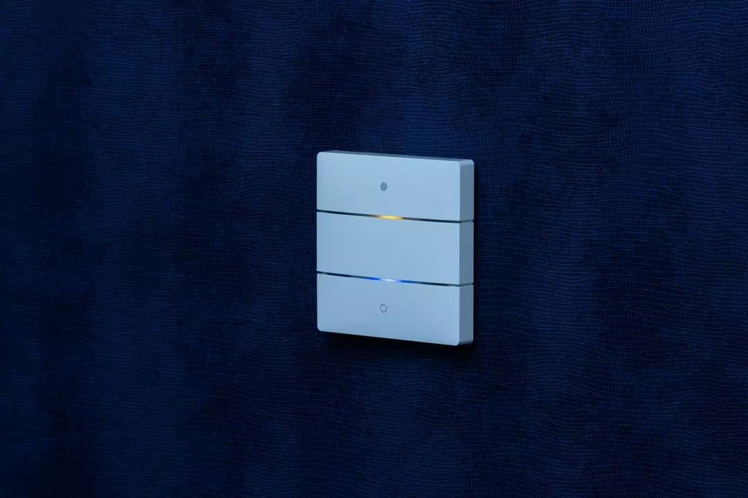 Ein moderner, weißer Lichtschalter mit drei horizontalen Tasten an einer dunkelblauen Wand. Eine Taste leuchtet schwach.