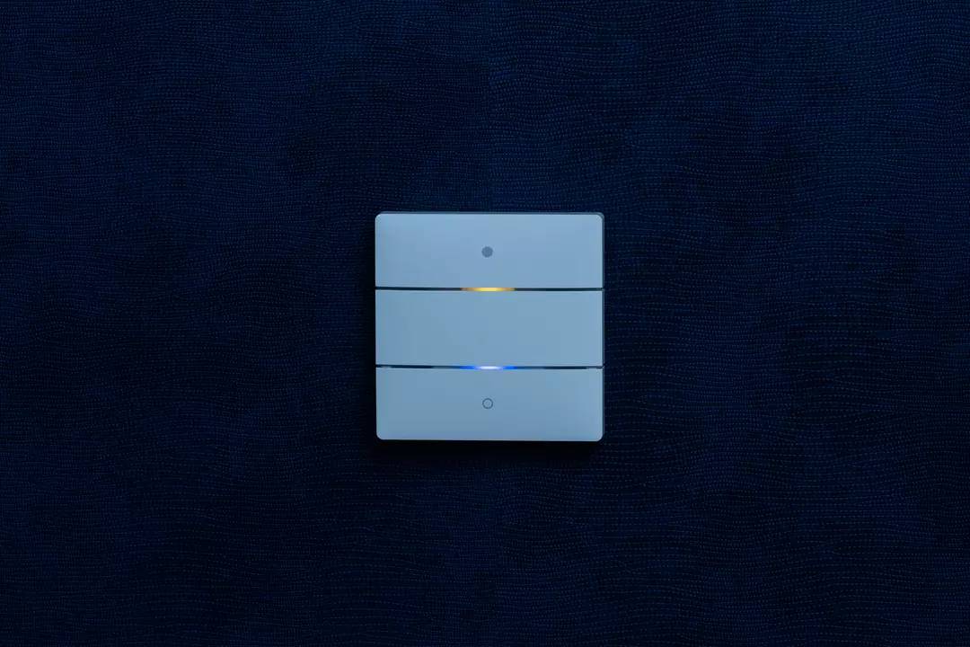 Ein dreifacher Lichtschalter an einer blauen Wand mit einer gelben und blauen Kontrollleuchte an zwei der Schalter.