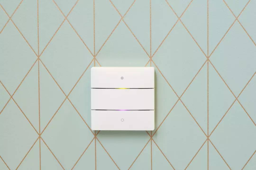 Ein weißer Lichtschalter an einer türkisfarbenen Wand mit goldenen Rautenlinien. Der Schalter hat drei horizontal geteilte Segmente.
